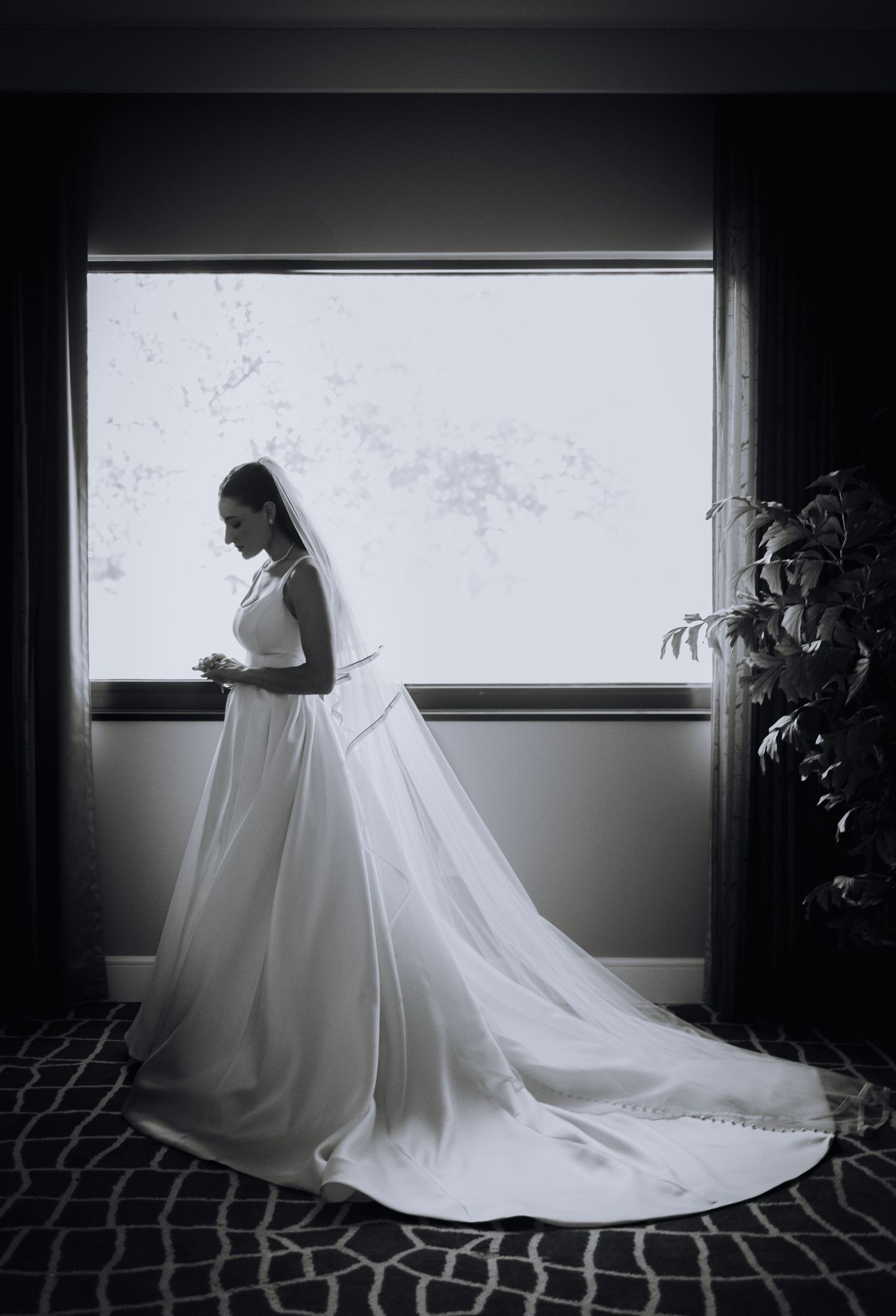 A bride in a flowing white wedding gown with long train stands silhouetted by a window in dramatic black and white.