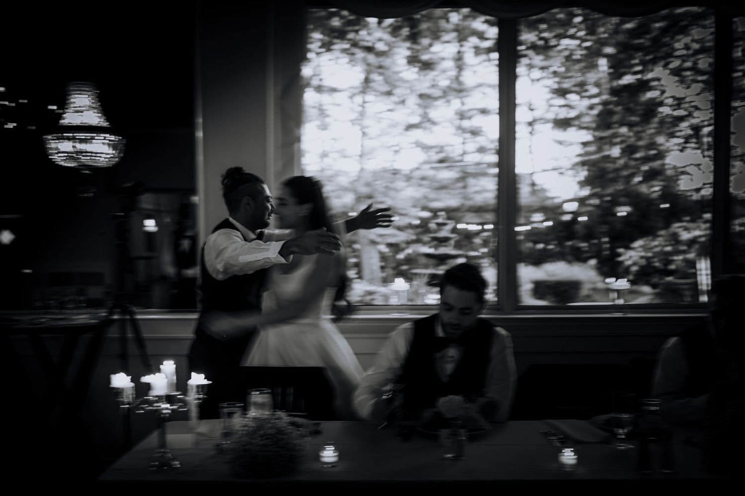 Black and white photo of couples dancing in an intimate candlelit setting with window views.