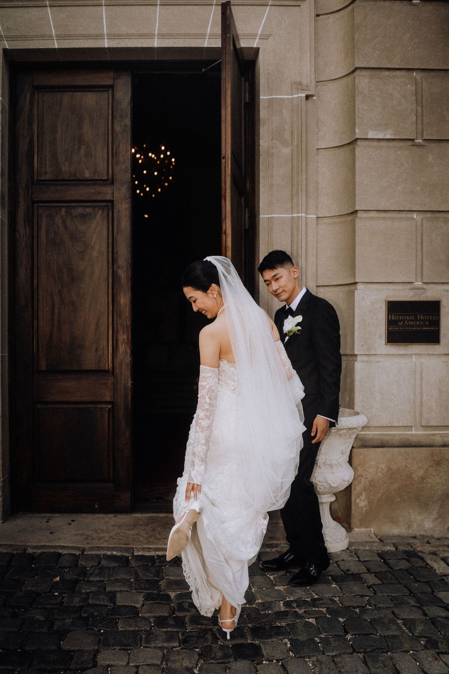 Asian bride in lace gown and veil with groom in tuxedo exiting elegant stone building on cobblestone street.