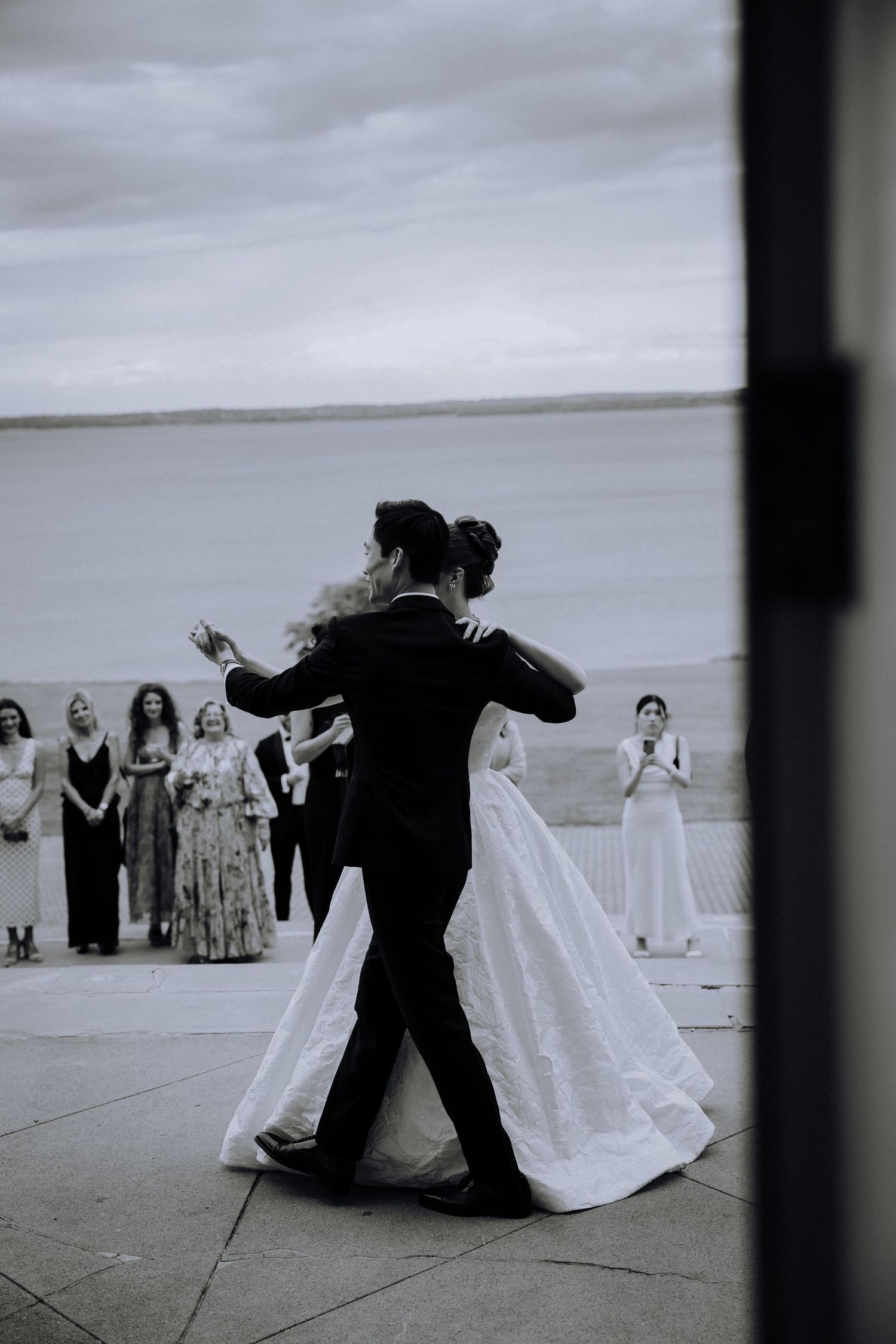 Black and white photo of bride and groom sharing first dance outdoors by waterfront with guests watching.