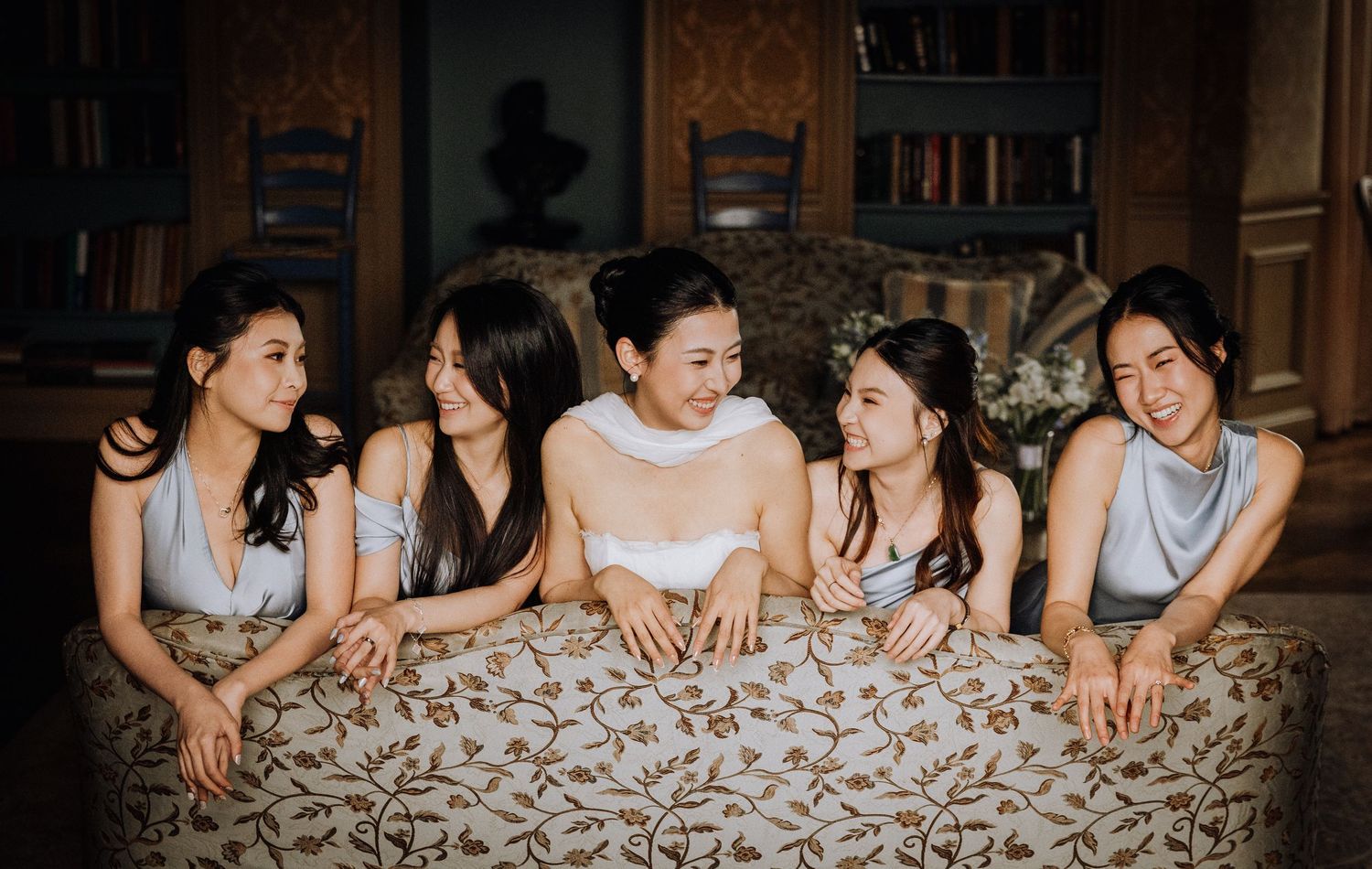 Bride in white dress poses with four bridesmaids in silver gowns, leaning over a floral couch in an elegant room.