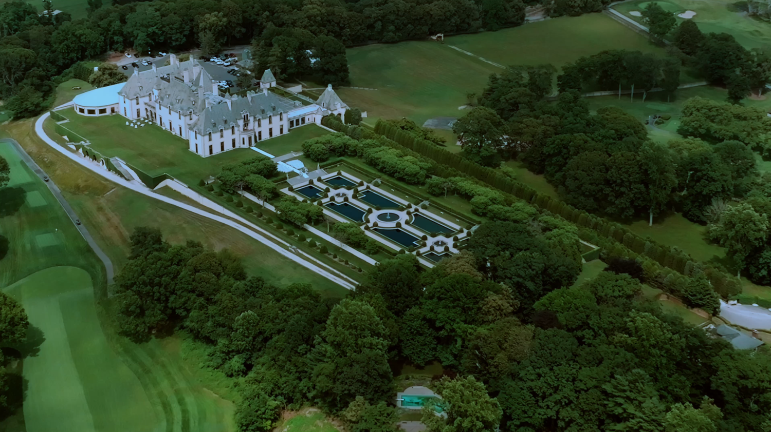 Aerial view of a grand estate mansion with manicured gardens, pool, and lush green surroundings.