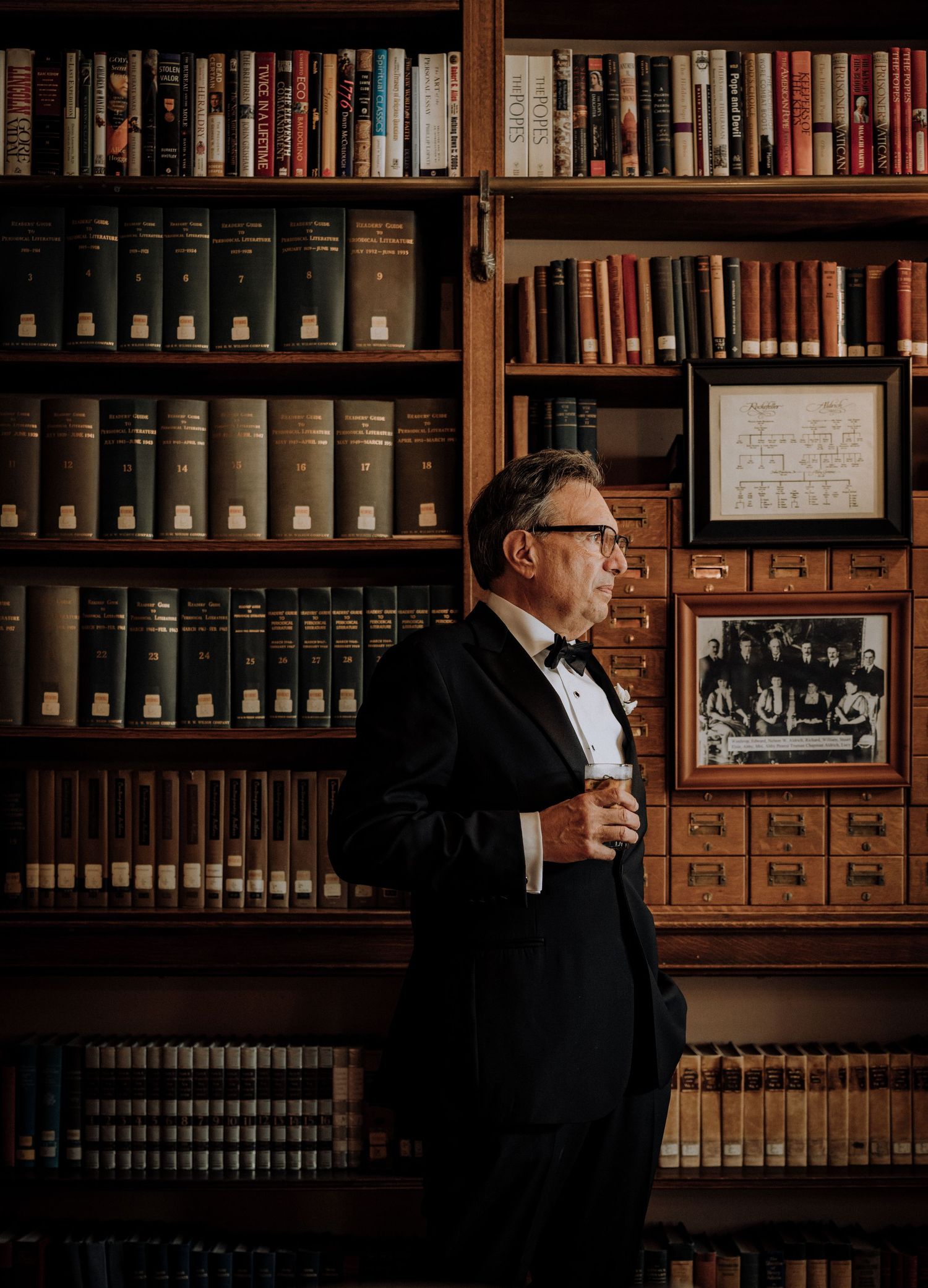 Distinguished man in tuxedo and bow tie stands in elegant library with floor-to-ceiling bookshelves.