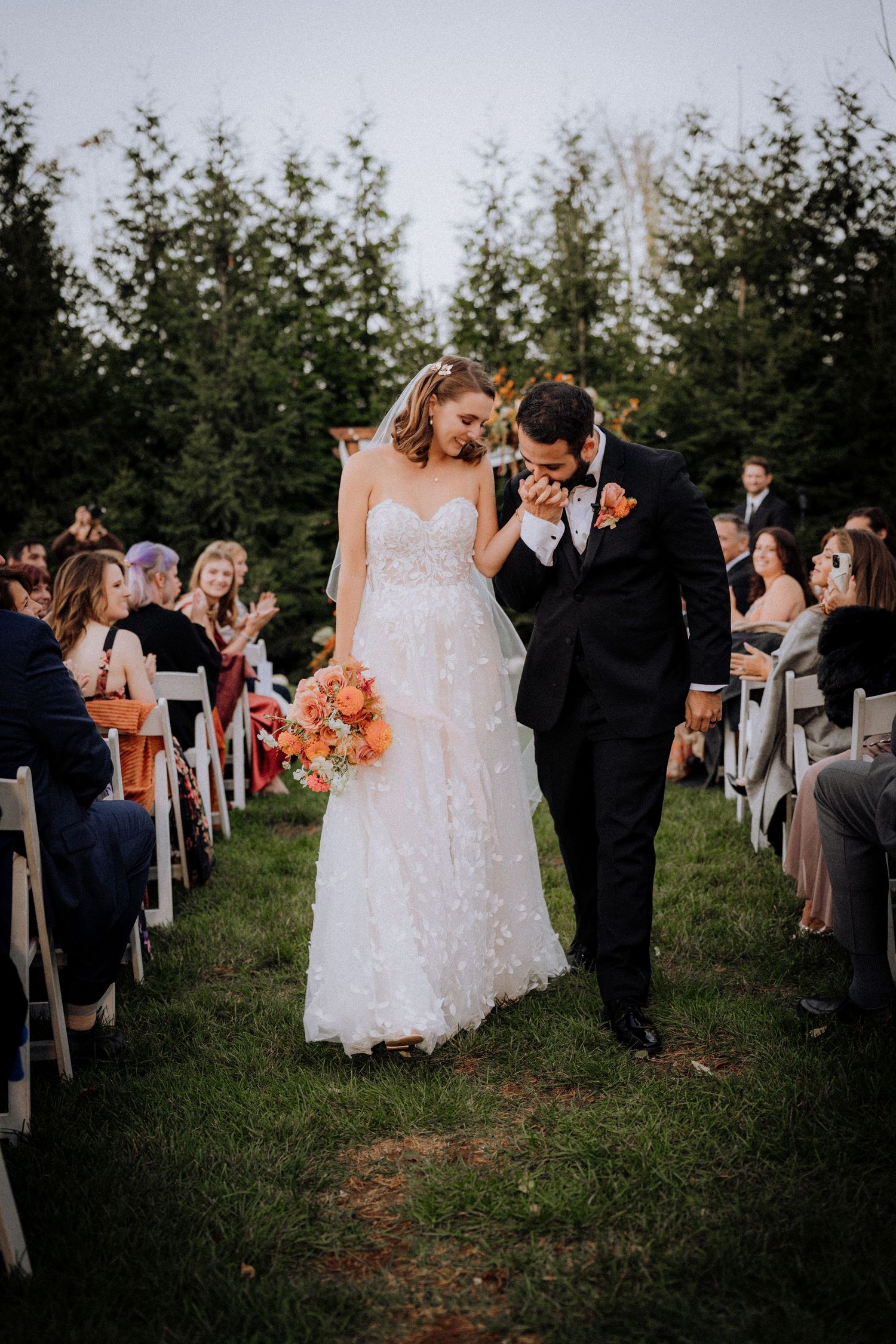 A newlywed couple walking down the aisle while guests celebrate at an outdoor ceremony.