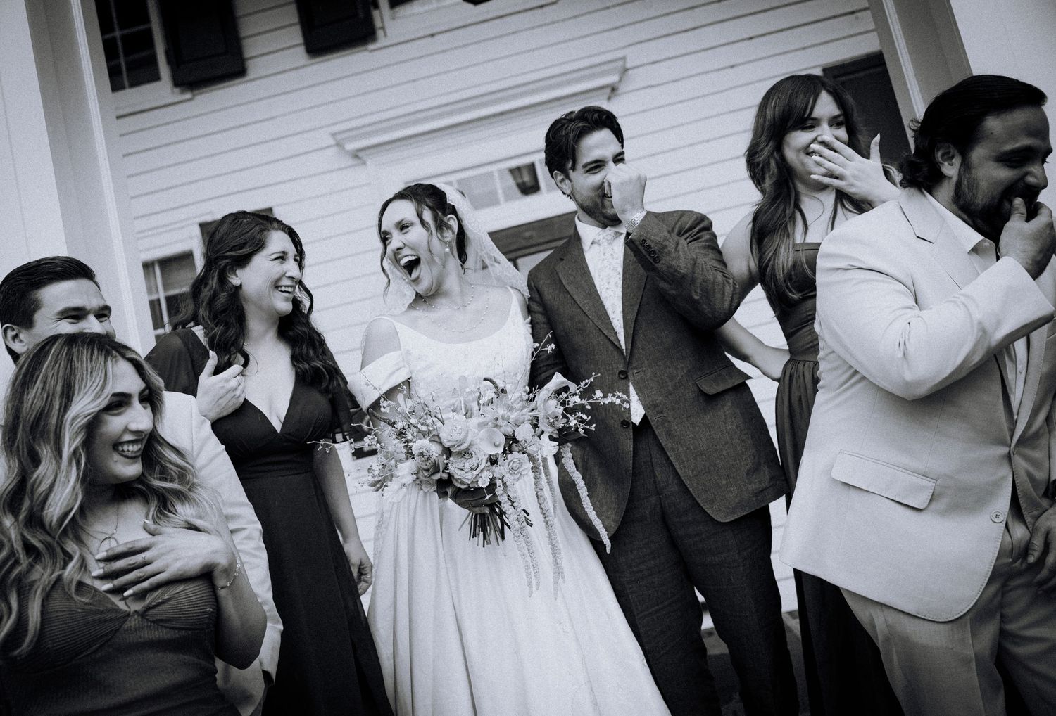Black and white wedding photo of bride and groom laughing with bridesmaids and groomsmen outside white building.