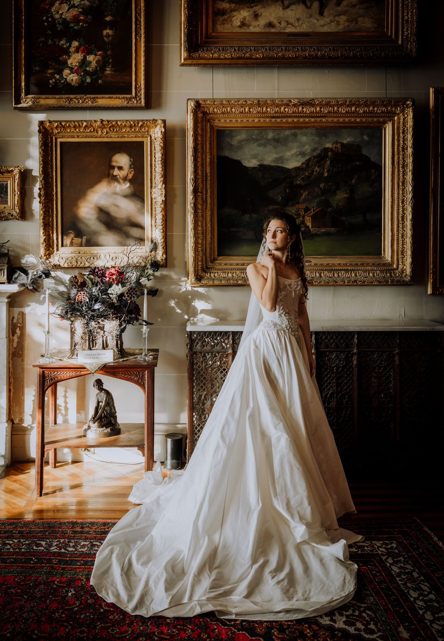 Bride in a flowing white gown with a long train stands in an ornate gallery with gilded framed paintings and a red patterned rug.
