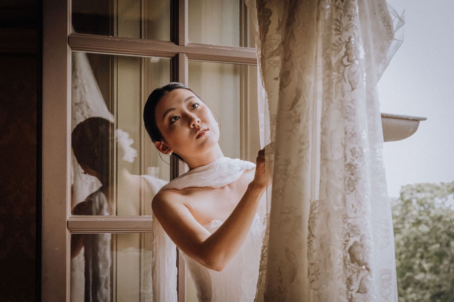 A young woman in a white dress gazes upward while holding lace curtains near a sunlit window.
