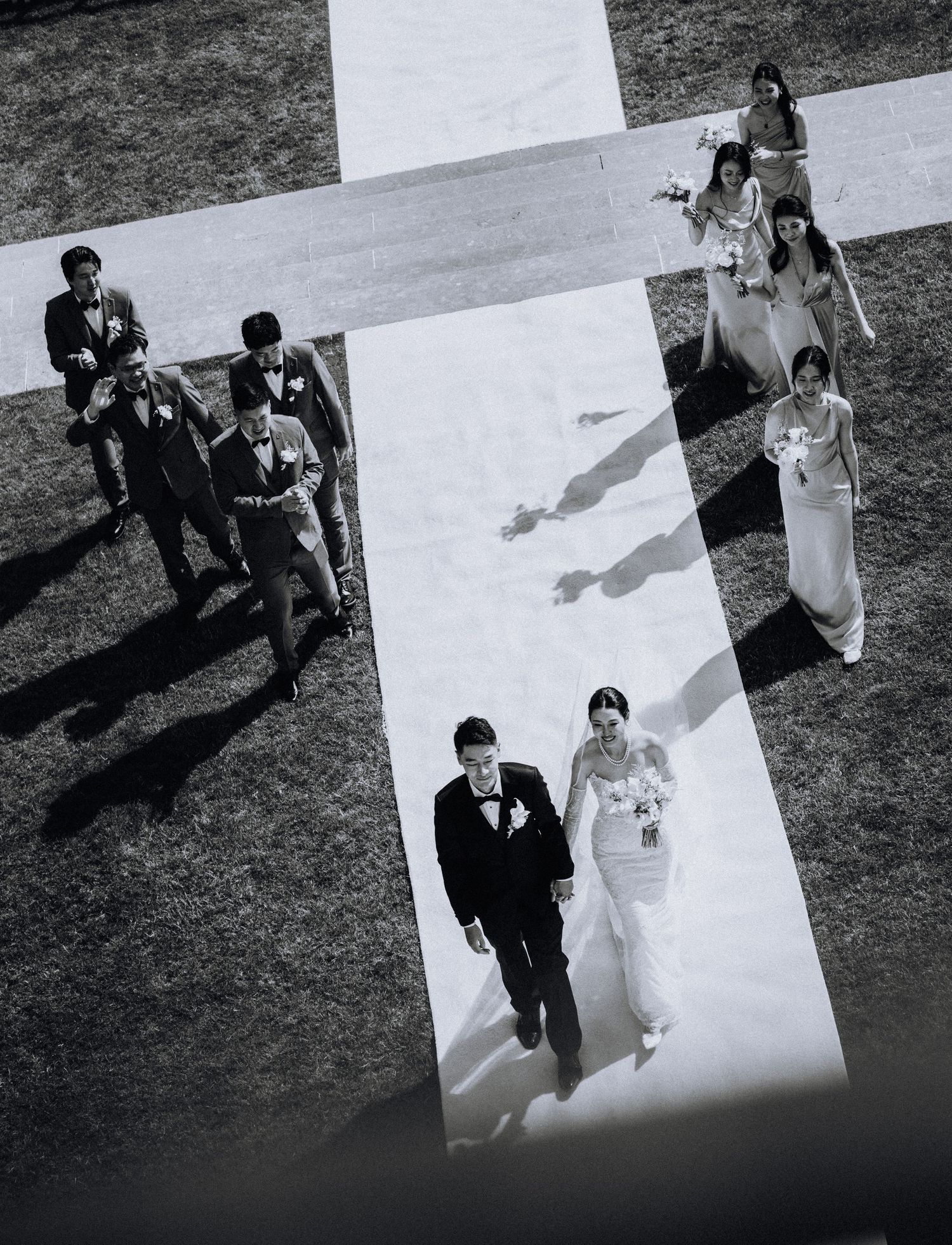 Aerial black and white photo of a bride and groom walking down an outdoor aisle with wedding party following behind.