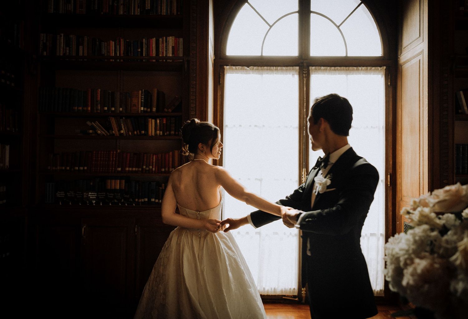 A bride and groom hold hands in a dimly lit library with tall bookshelves and an arched window backdrop.