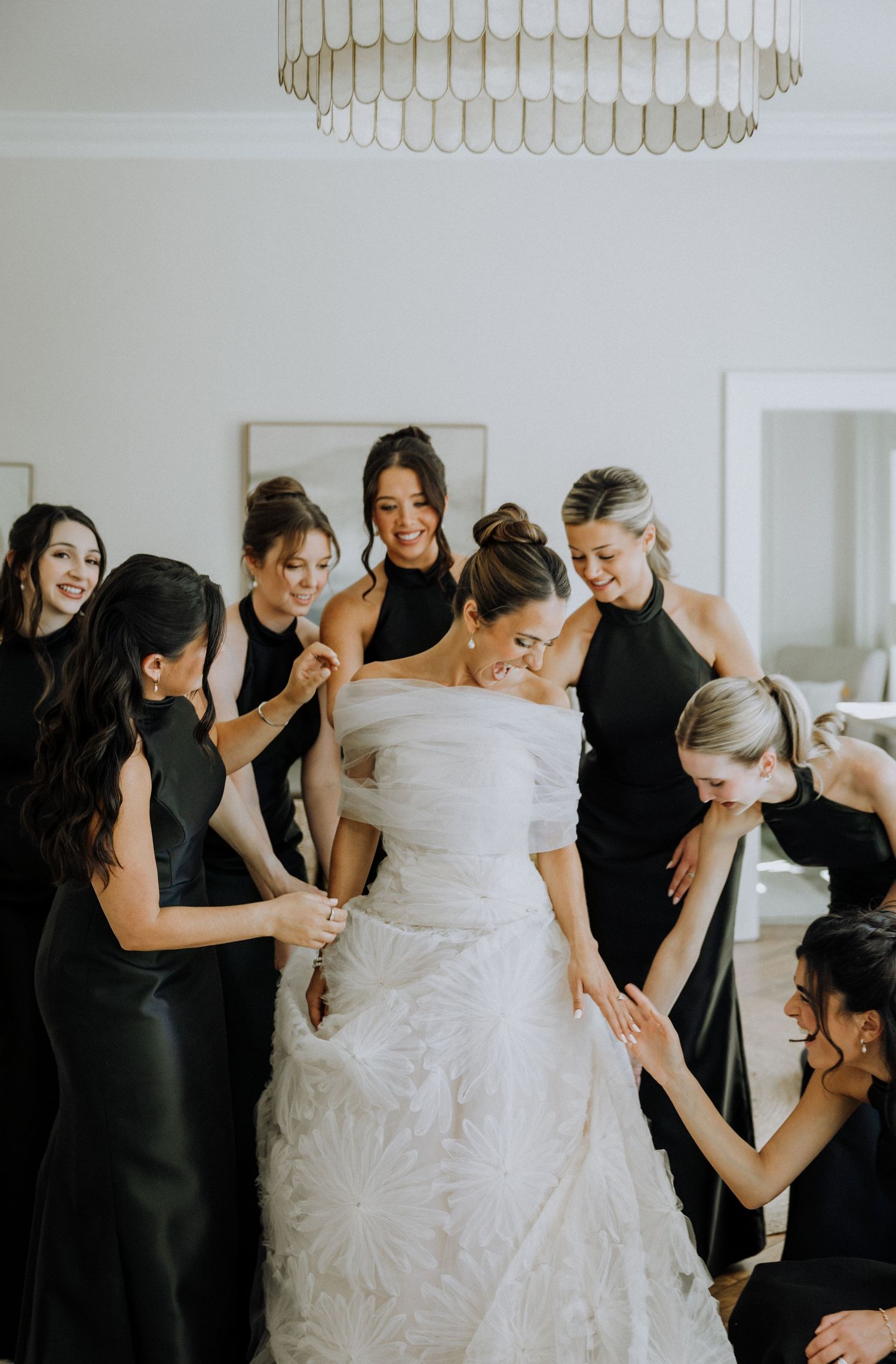 Bride in white lace gown surrounded by bridesmaids in black dresses helping her get ready indoors.