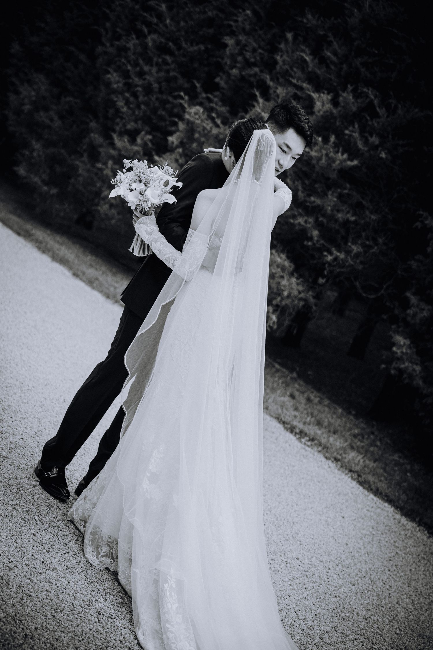 Black and white photo of a bride and groom sharing a romantic kiss, with flowing veil and bouquet outdoors.