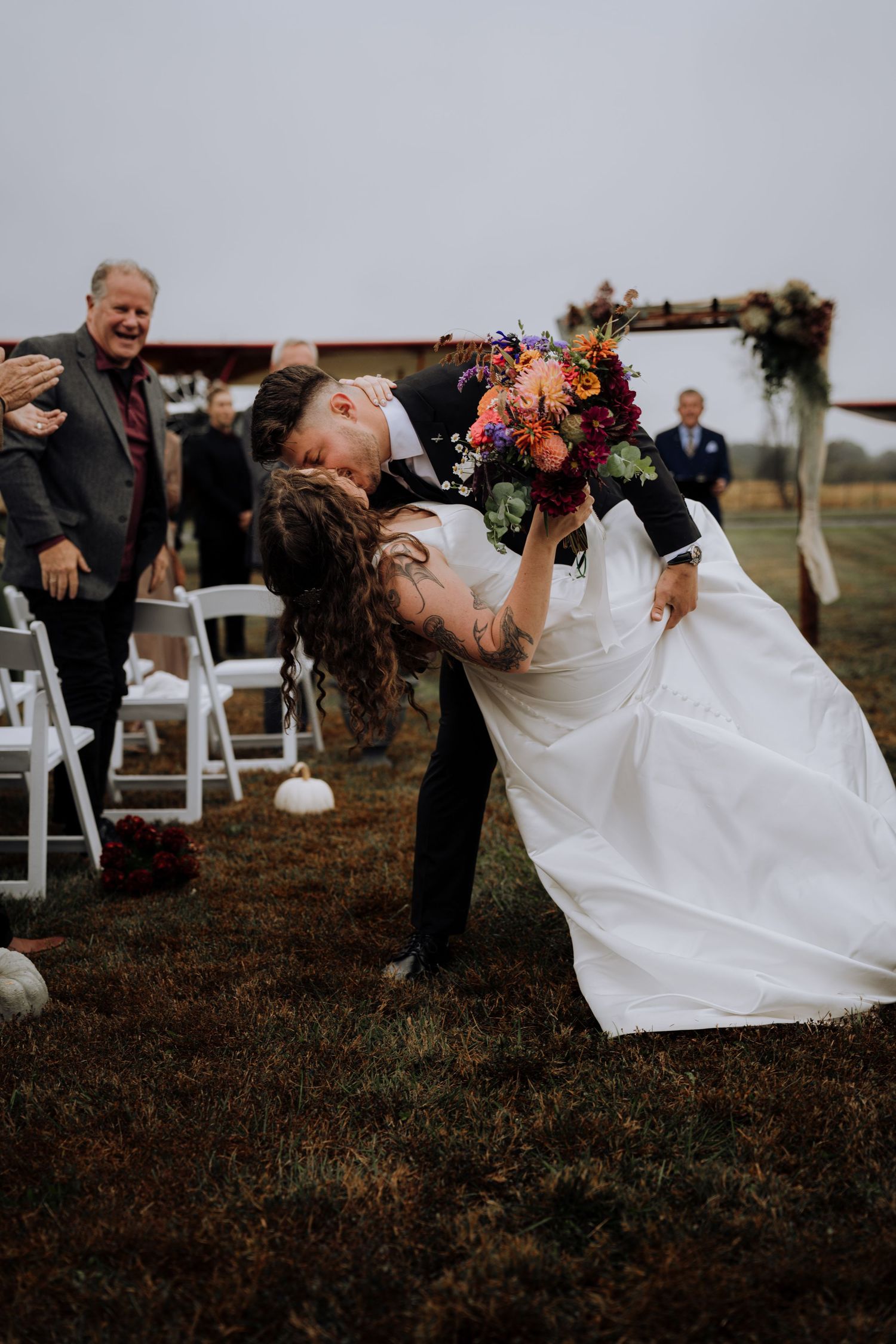 A bride in a white dress performs a dramatic dip pose during an outdoor wedding ceremony.