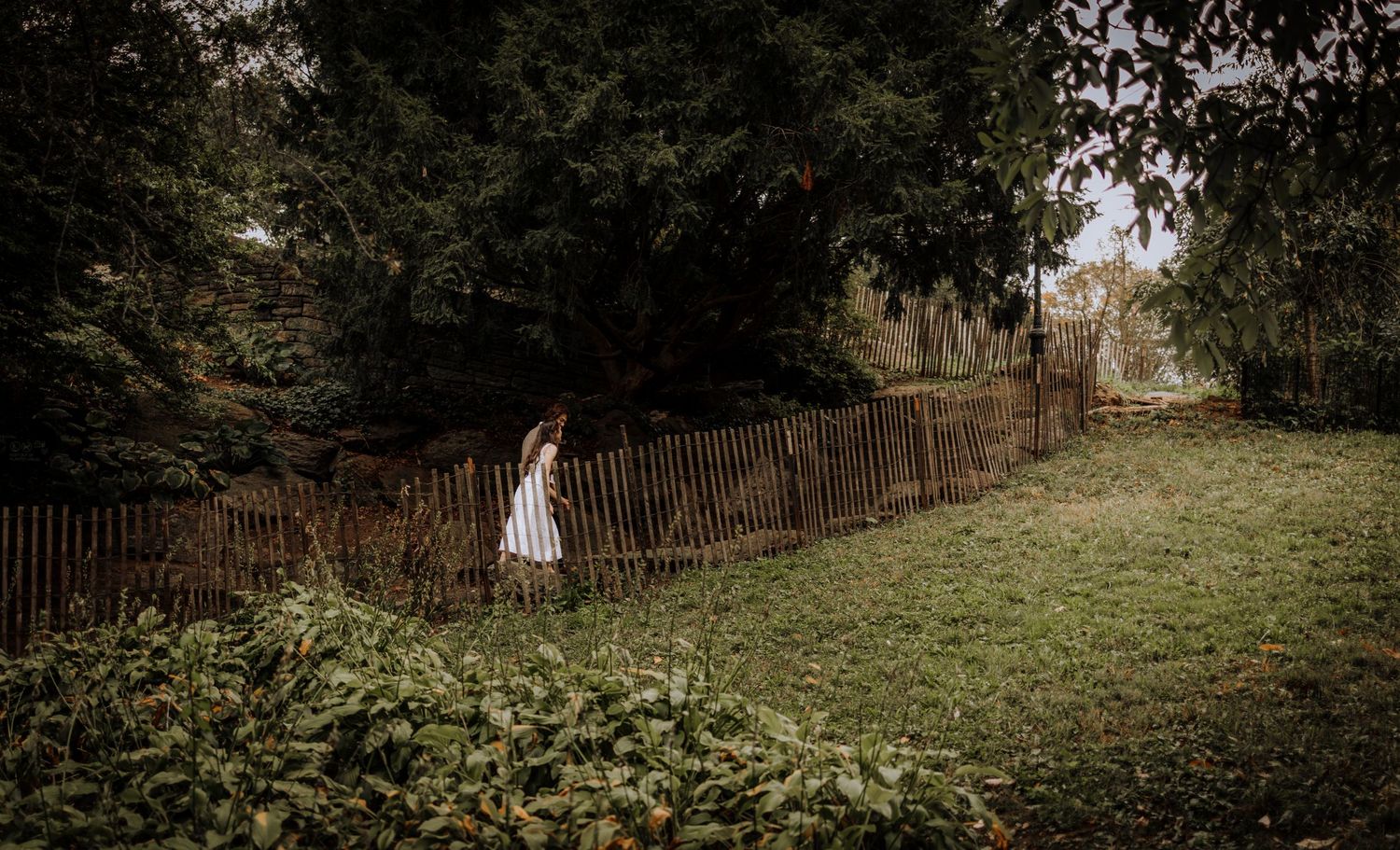 A solitary figure in white stands by a rustic fence in a misty countryside setting.