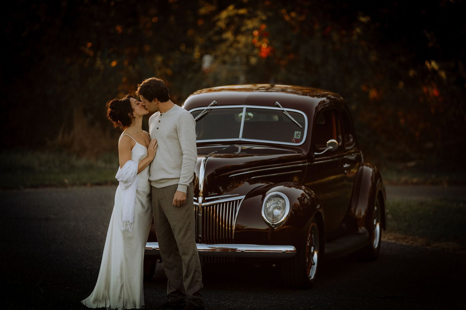 Bride in a flowing white gown kisses her groom beside a vintage black car at dusk, with autumn foliage glowing in the background.