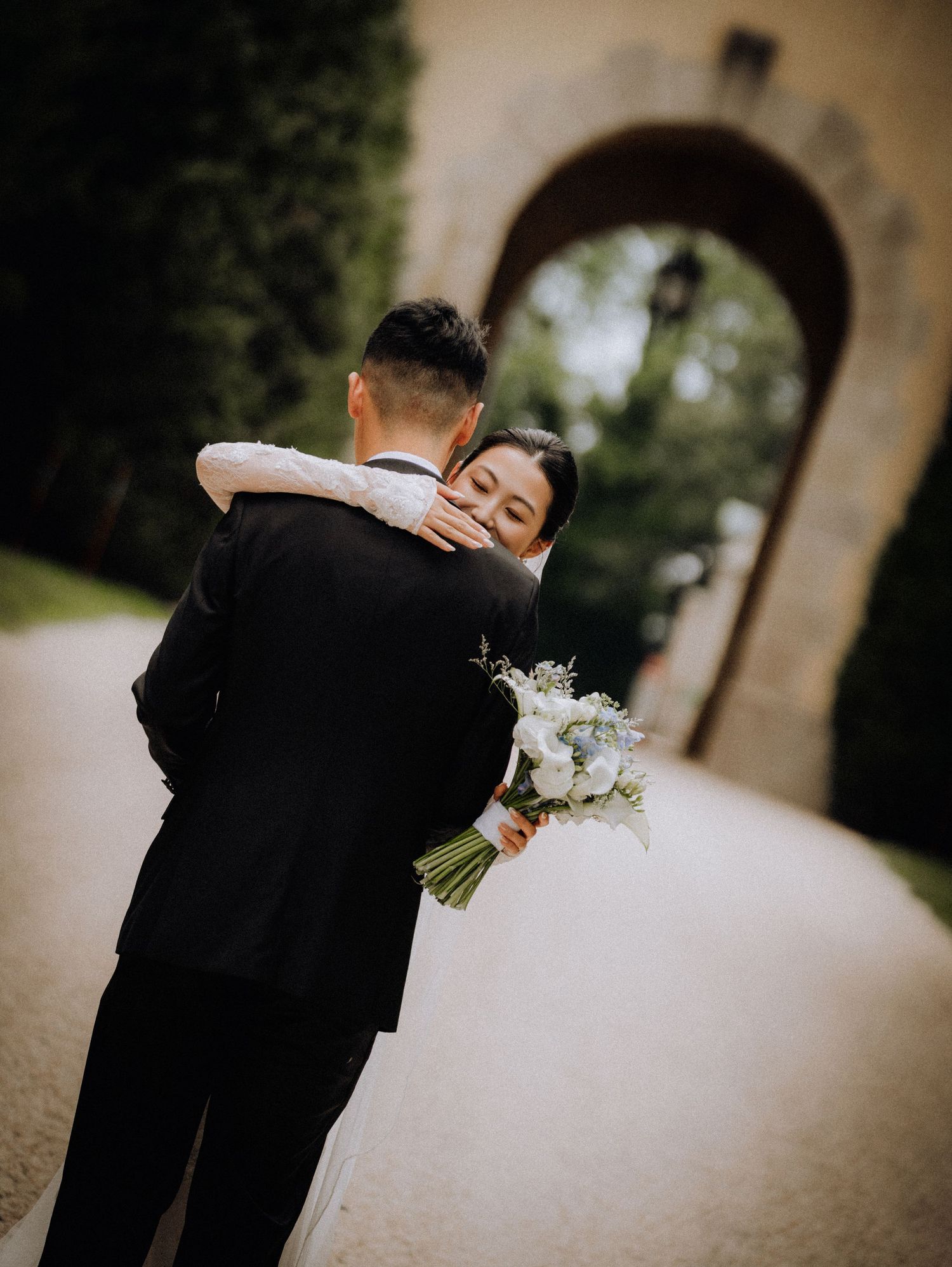 Bride holding white bouquet embraces groom in black tuxedo on elegant pathway with stone arch backdrop.