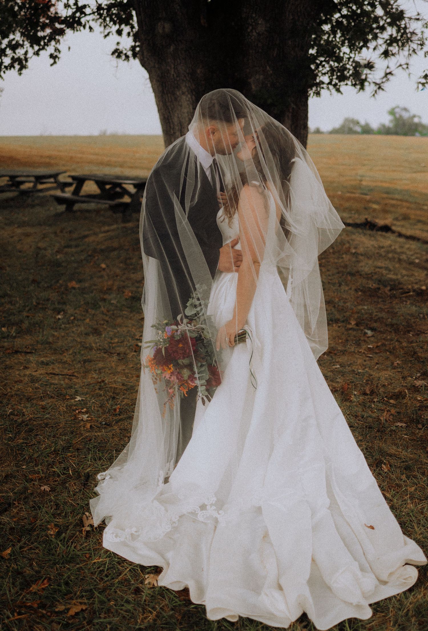 A bride in a flowing white dress and veil stands gracefully in an autumn field at sunset.