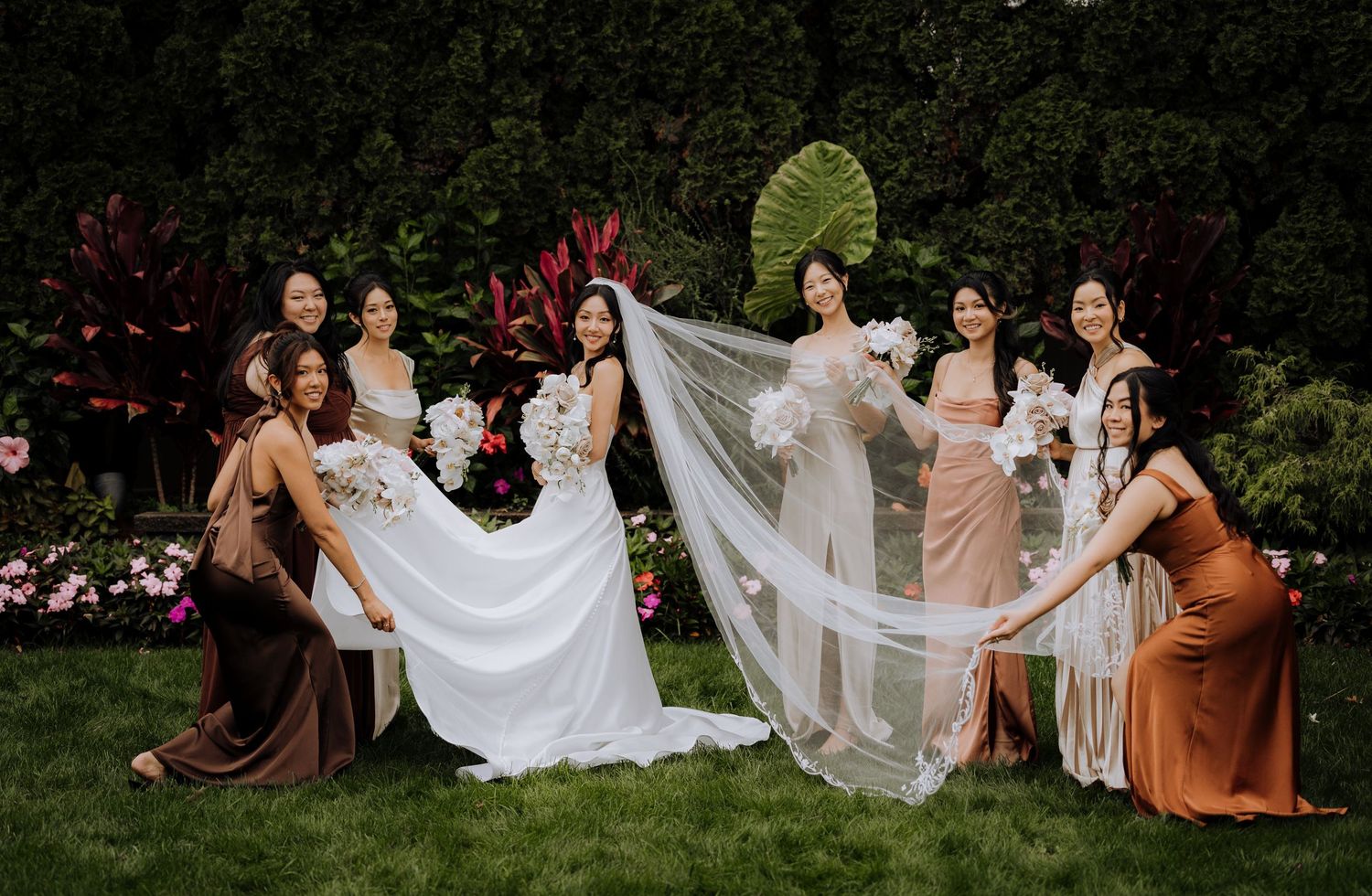 Wedding party sits on grass holding flowing white dress fabric in garden setting.