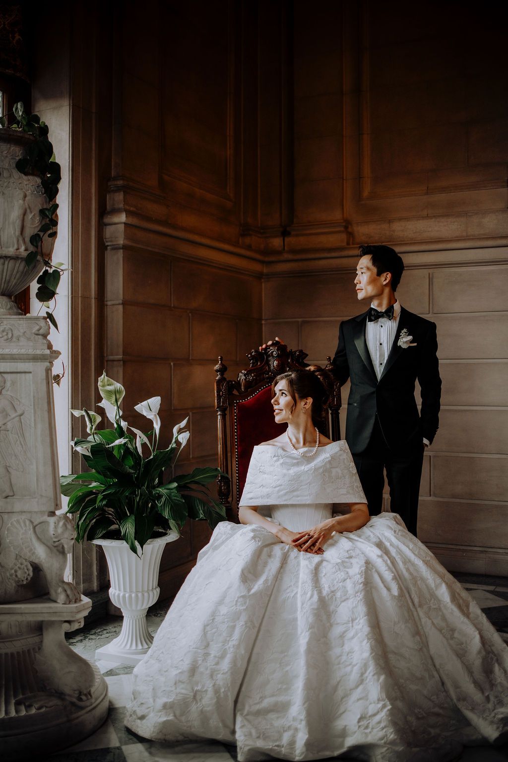 Elegant bride in white ball gown sits beside groom in black tuxedo in an ornate wood-paneled room with white calla lilies.