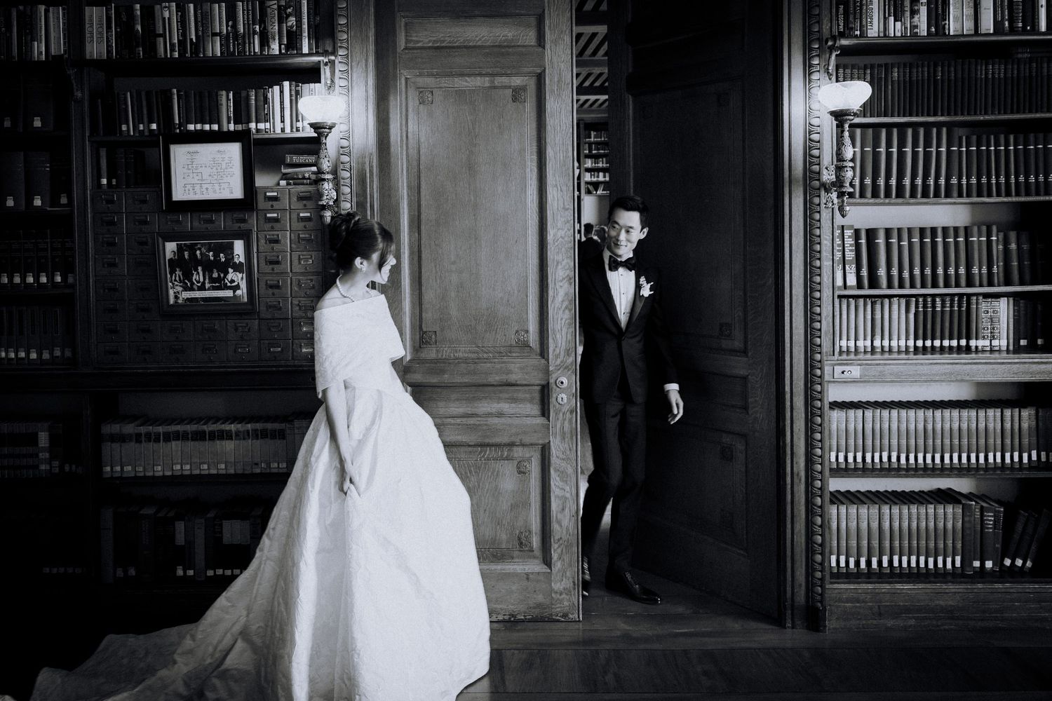 Black and white wedding photo of bride and groom in elegant library with wood paneling and bookshelves.