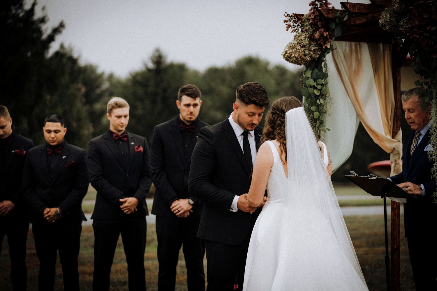 Outdoor wedding ceremony with wedding party in black attire standing before wooden arch.