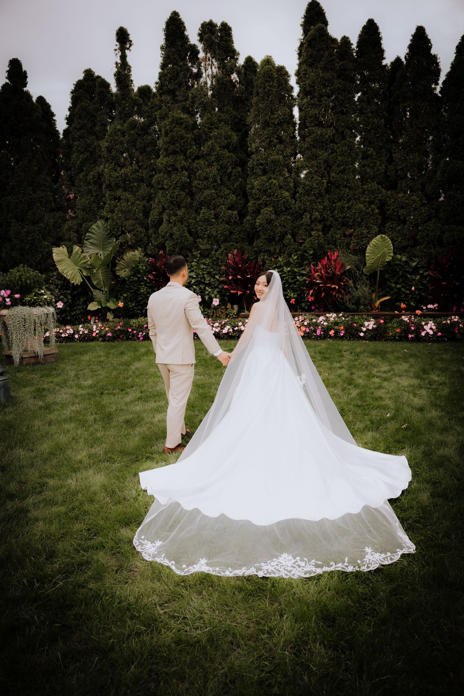Wedding couple walking through a garden with cypress trees as the bride's white dress trails behind them.