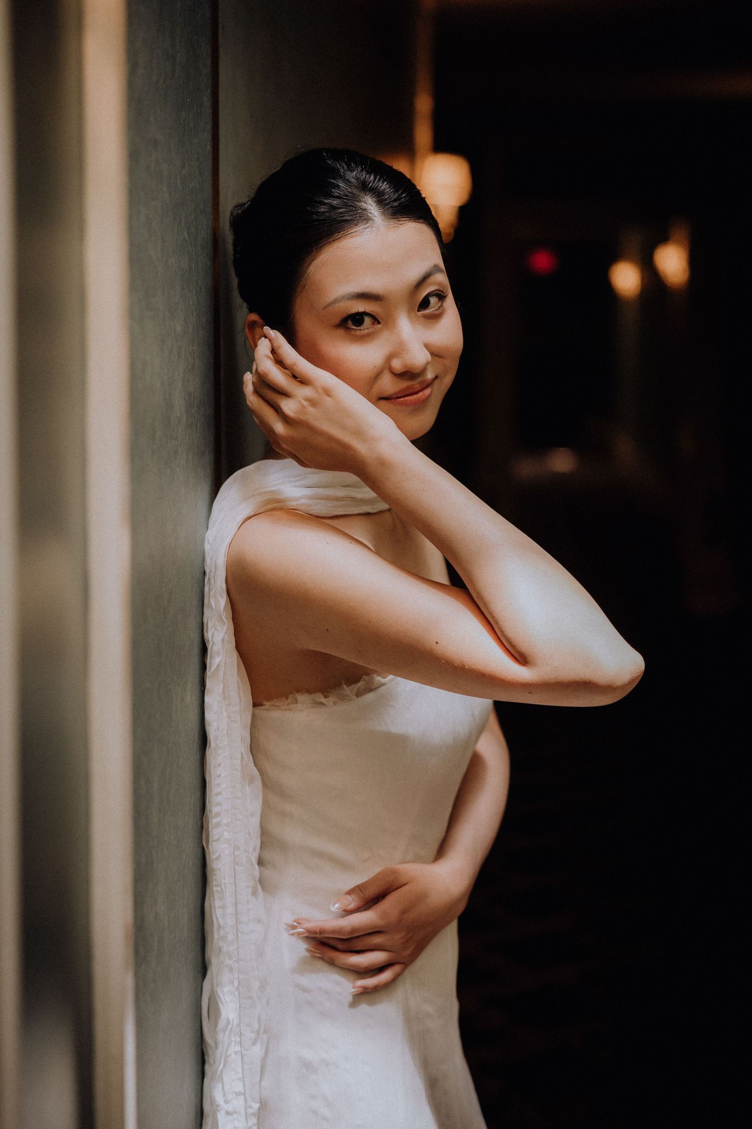 Elegant Asian bride in white gown leaning against a column, smiling softly in a dimly lit venue.