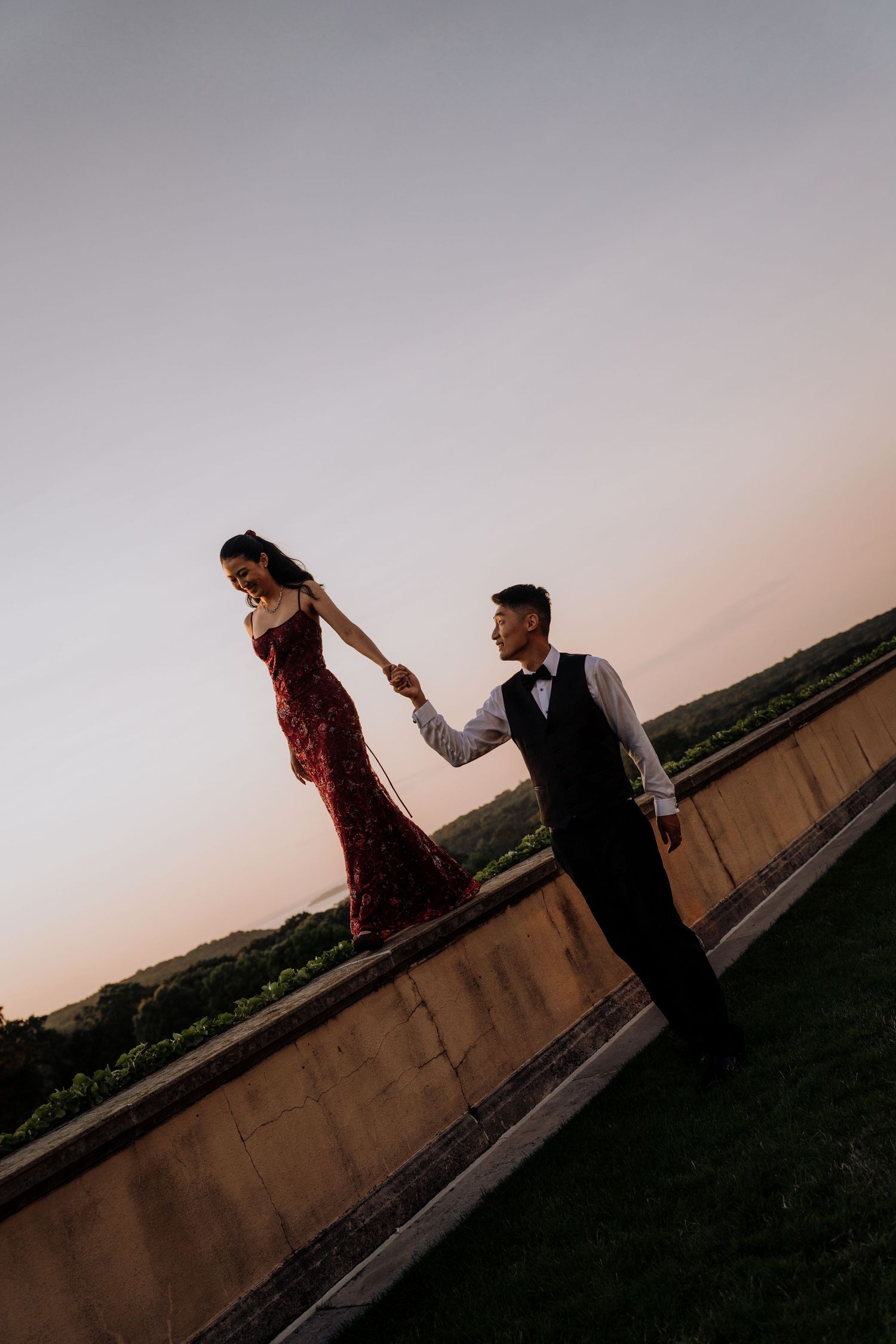 Couple in formal wear holding hands at sunset, woman in red sequin gown standing on wall, man in black tuxedo.