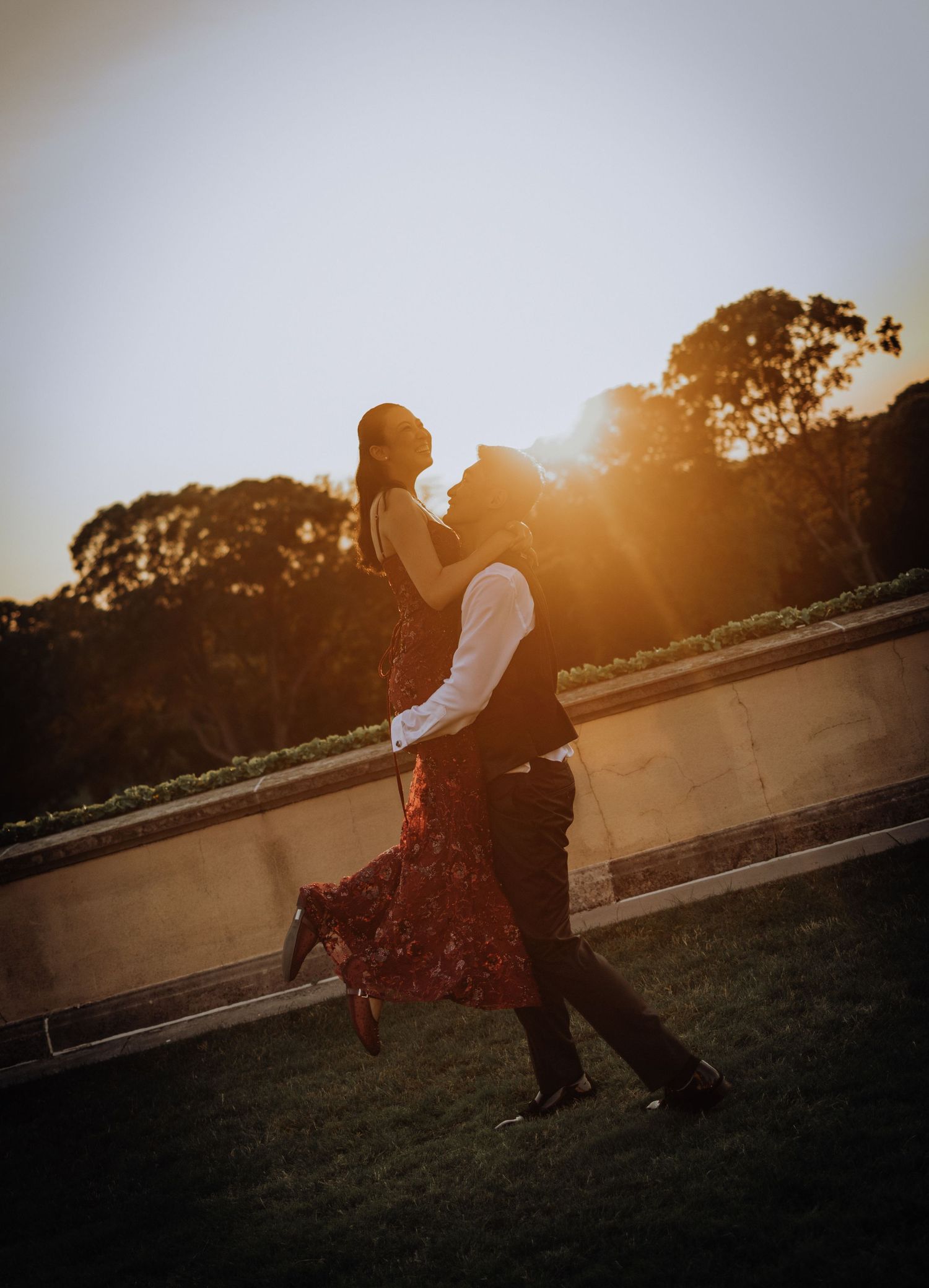 Couple dancing romantically at golden hour sunset, silhouetted against warm glowing light with trees in background.