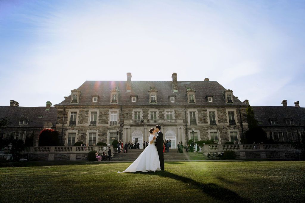 Bride and groom share a romantic kiss on lush green lawn before a grand French chateau at sunset.
