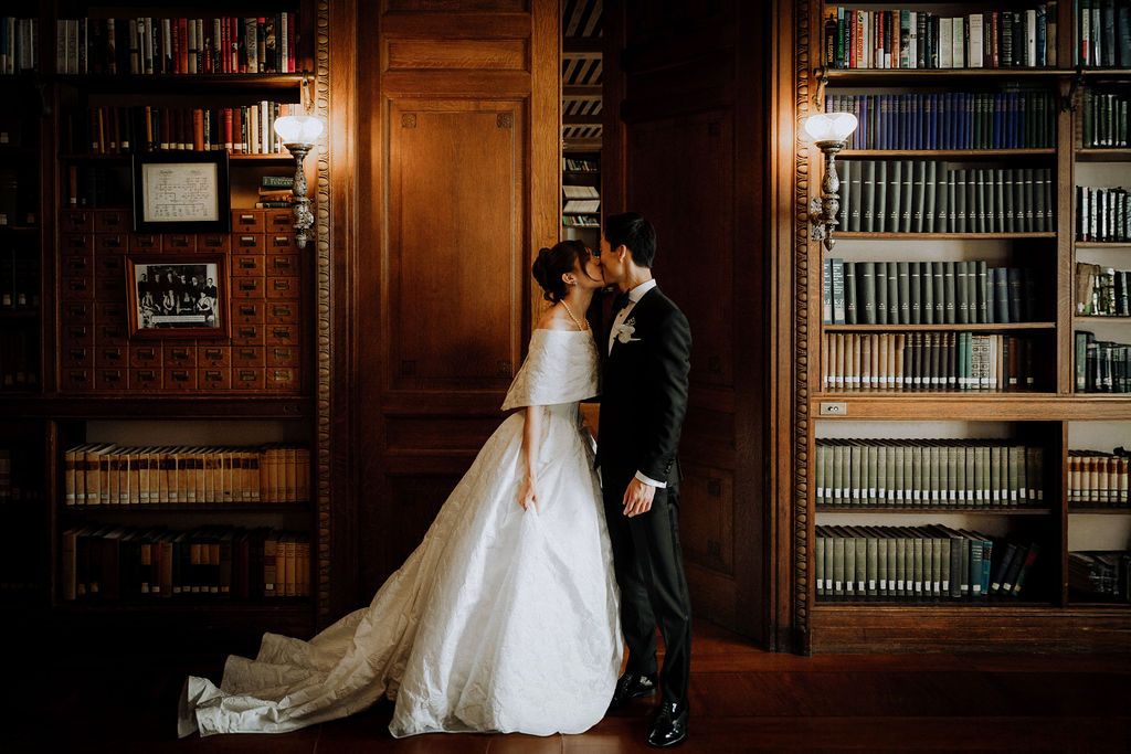 Bride and groom sharing a romantic kiss in an elegant wood-paneled library with floor-to-ceiling bookshelves.
