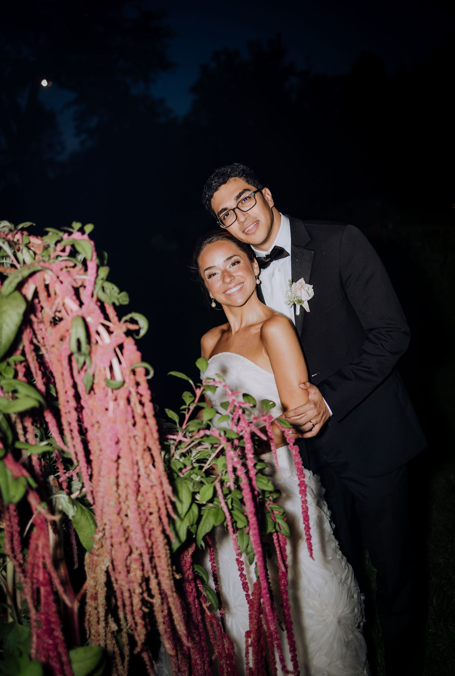 Bride and groom pose together at night beside cascading pink amaranth flowers during outdoor wedding celebration.