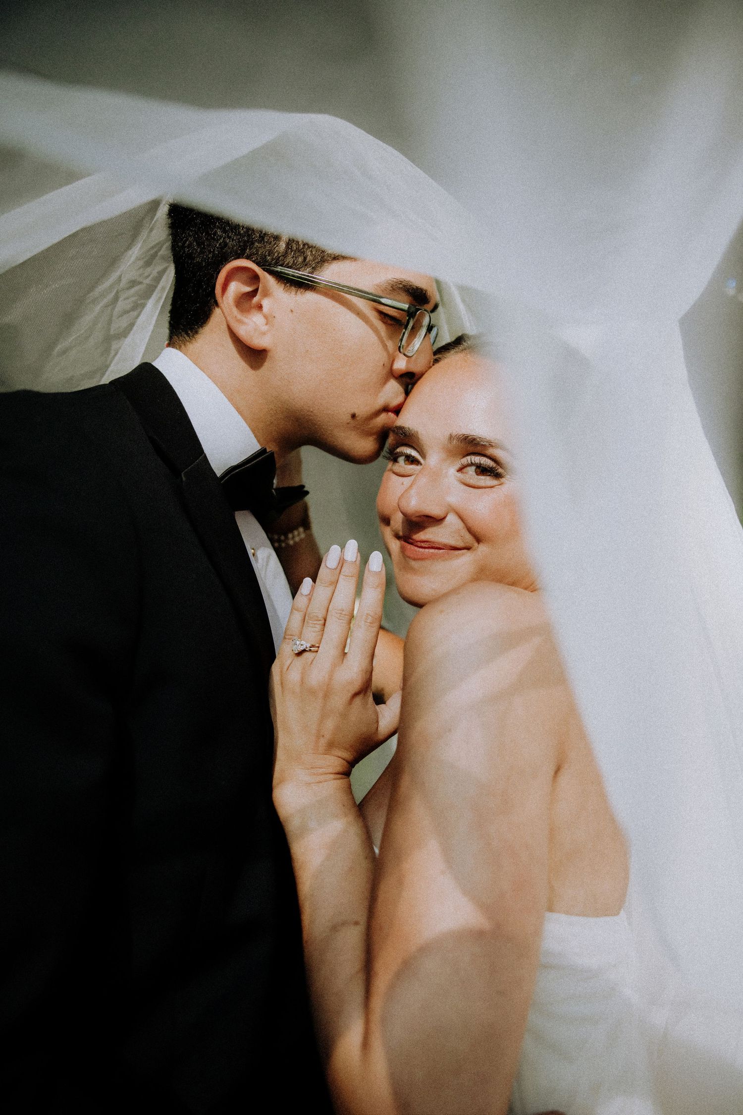 Groom kissing bride on cheek under wedding veil, bride smiling at camera wearing diamond ring.