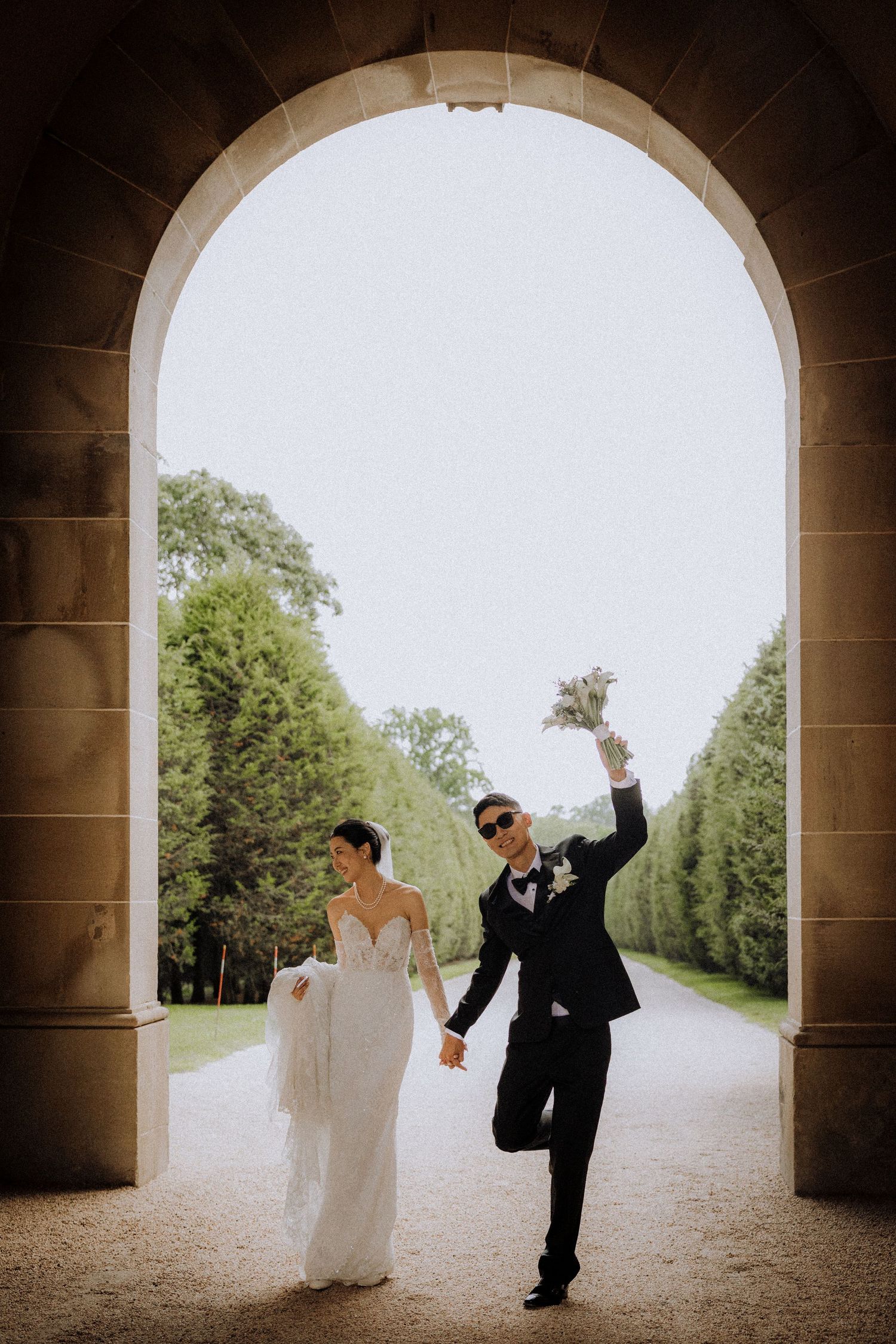 Bride and groom celebrate under stone arch, holding hands, groom raising bouquet, lush green garden path behind them.