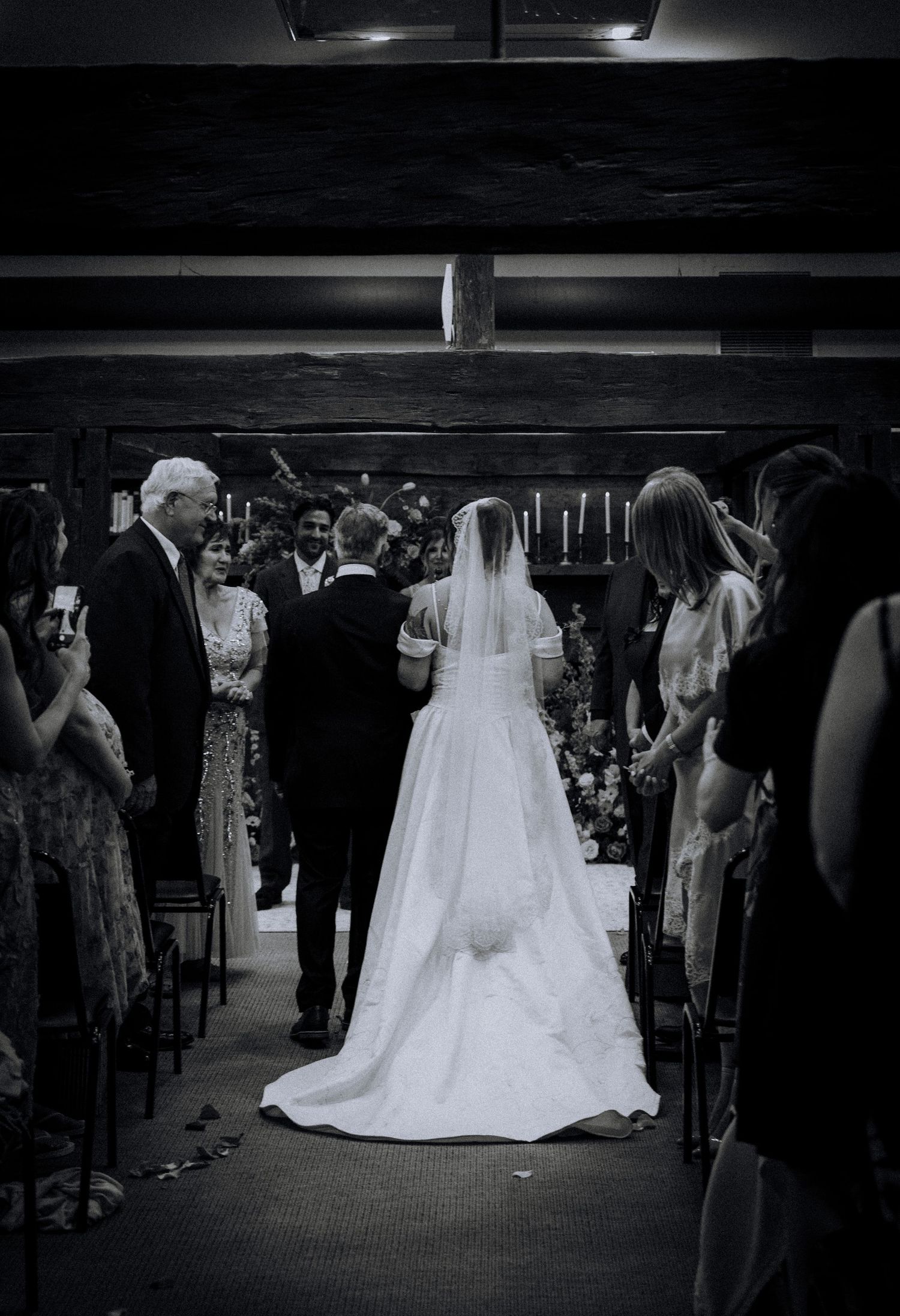 Black and white wedding ceremony photo showing bride in white gown and veil walking down the aisle with guests.