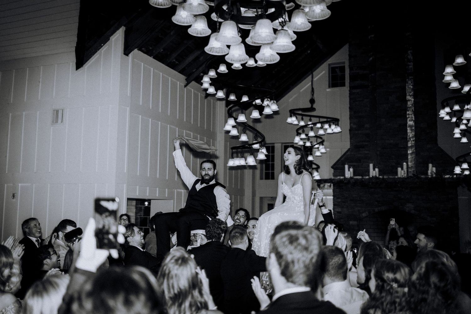 Black and white photo of a crowded wedding reception dance floor under pendant lights.