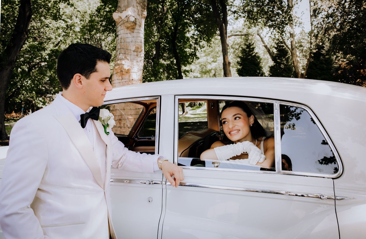 A classic car is featured in a wedding photoshoot with elegant white attire and natural outdoor scenery.