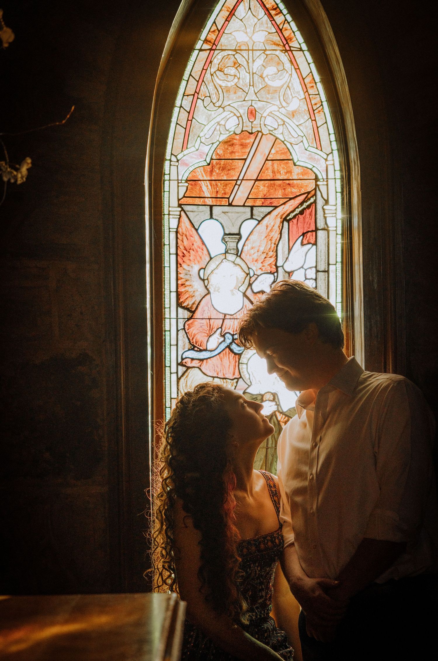 Couple embracing in front of a beautiful stained glass church window with orange and white patterns.