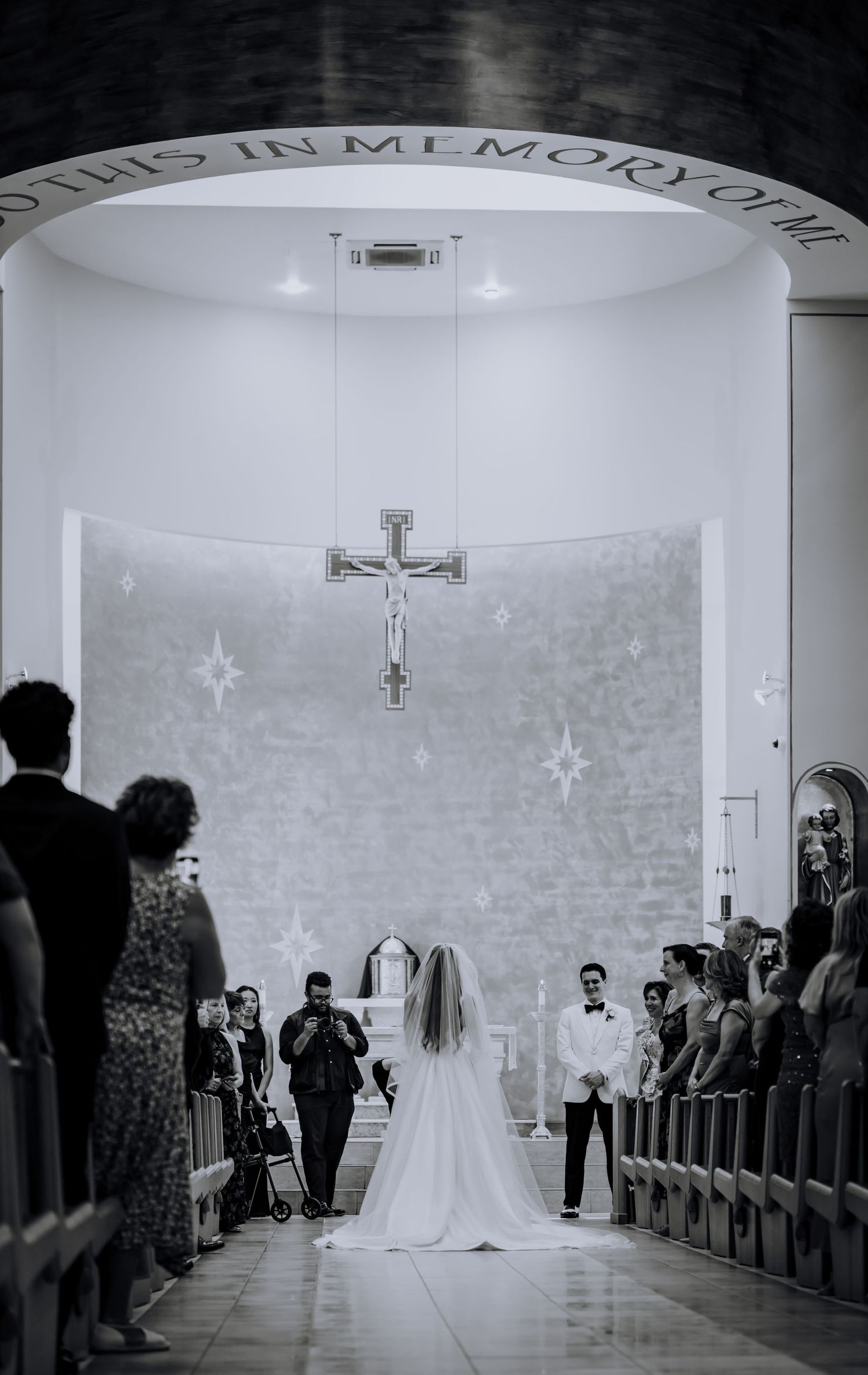 Black and white photo of a wedding ceremony in a minimalist church with curved white walls and crosses.Editorial and Cinematic Wedding photo
