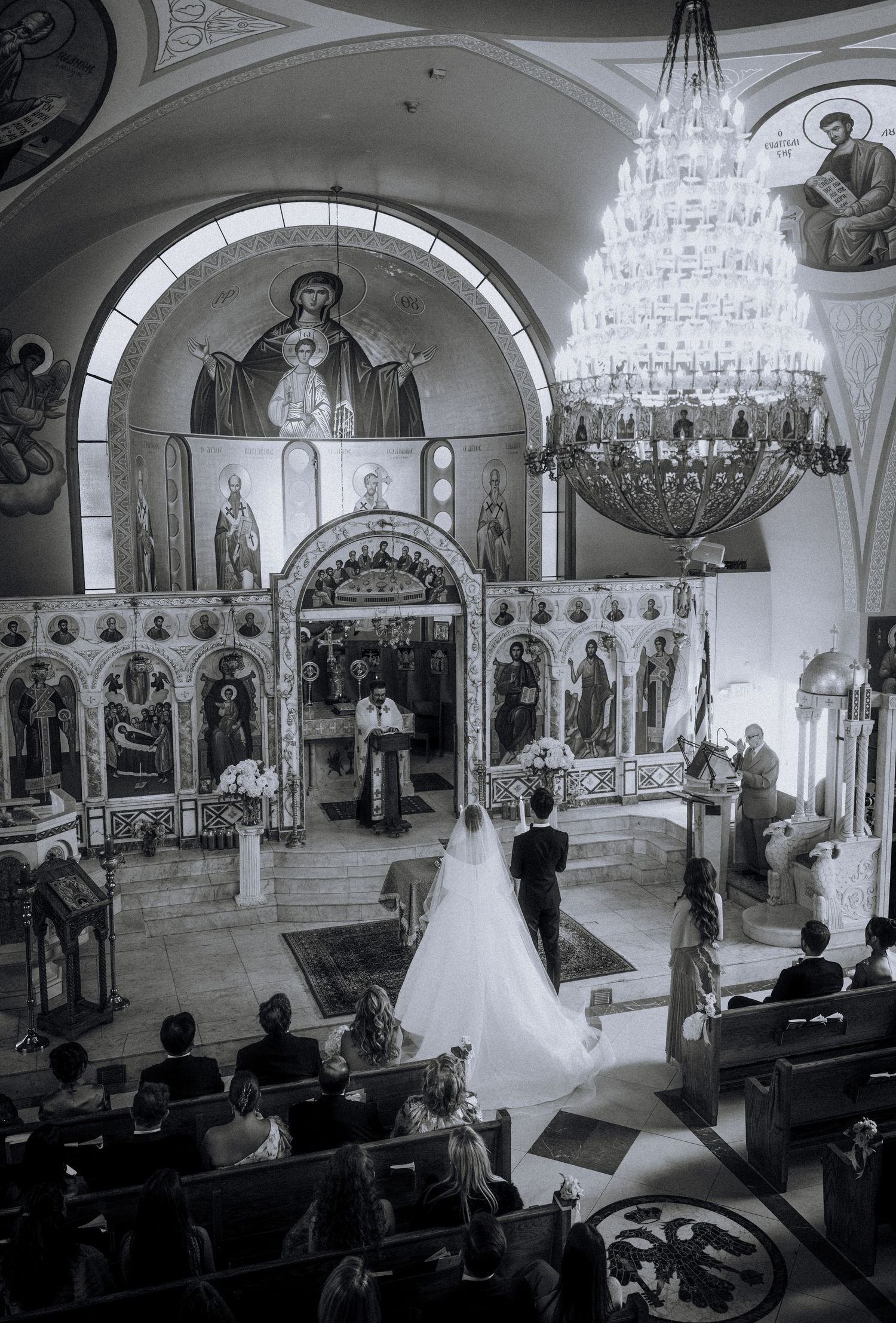 Black and white photo of a wedding ceremony inside an ornate Orthodox church with iconostasis and chandelier.