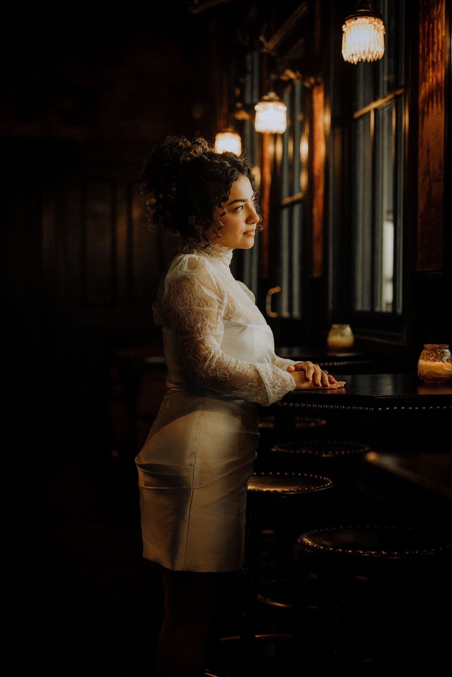 Woman in white lace dress standing at a dark bar, gazing thoughtfully toward a window in moody lighting.seven-grand-engagement-photos-los-an