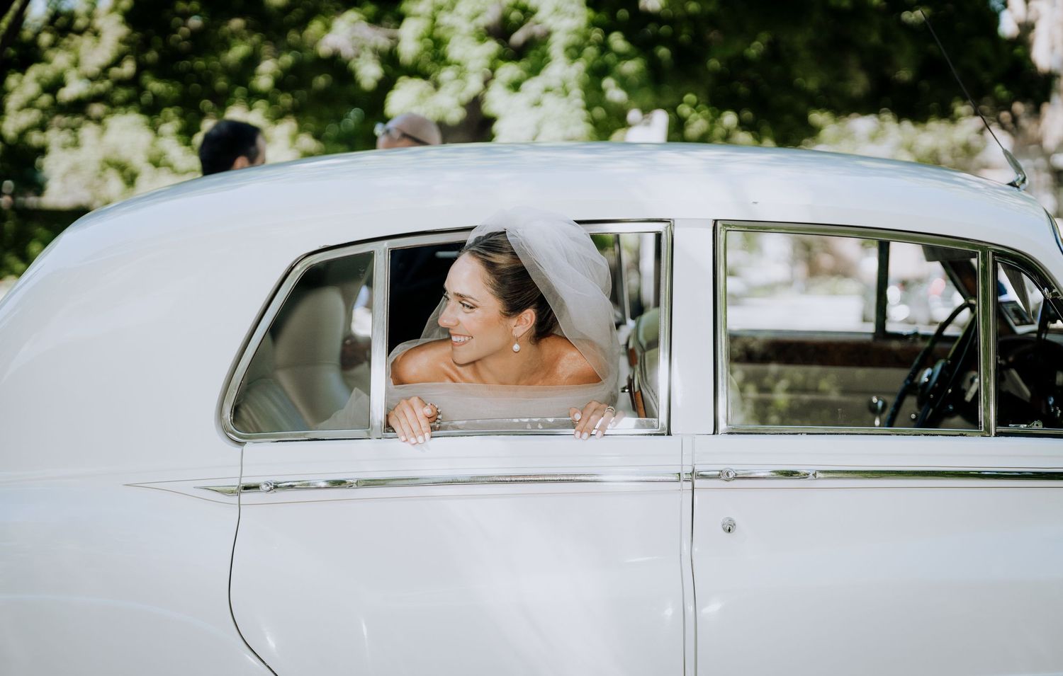 Bride in wedding veil smiling out of vintage white classic car window on her wedding day.