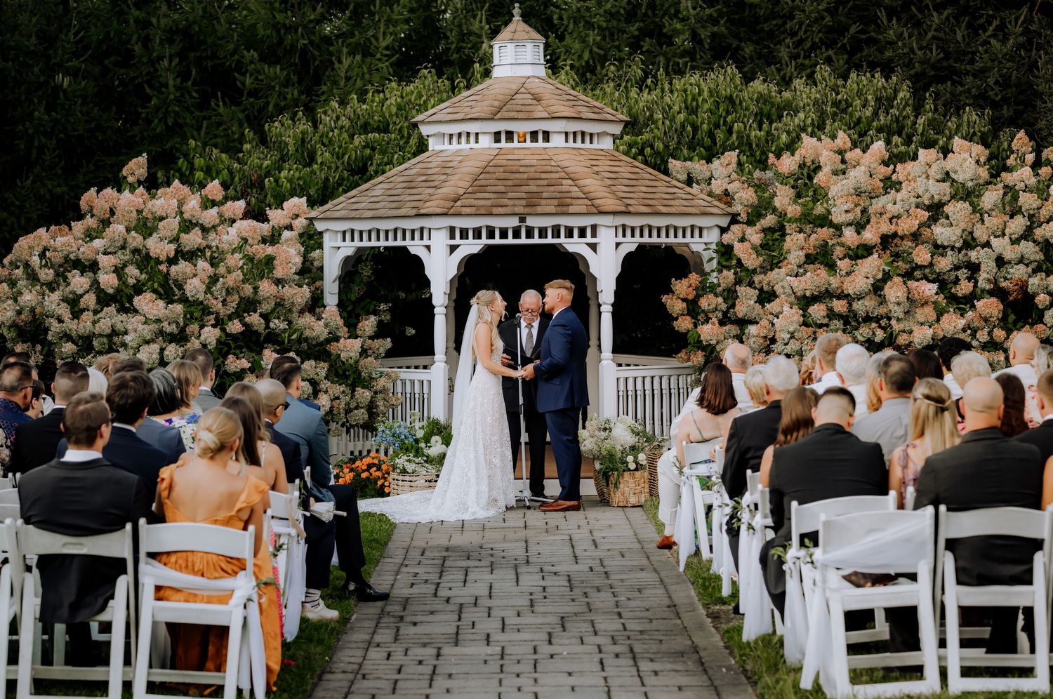 Wedding ceremony taking place at a white gazebo surrounded by blooming flowers and seated guests.Editorial and Cinematic Wedding photos