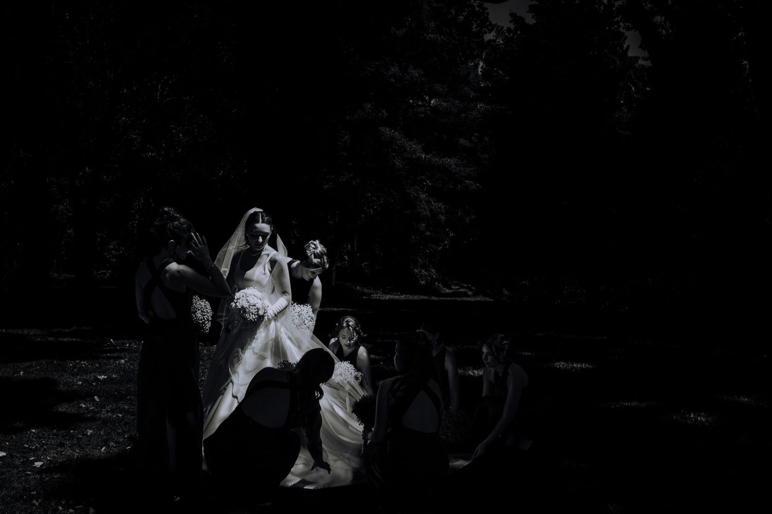 Black and white photo of a bride in white gown surrounded by attendants in dark outdoor wedding setting.Editorial and Cinematic Wedding phot