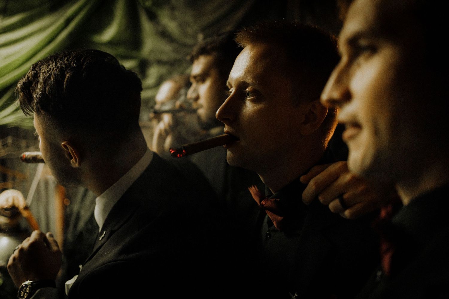 Wedding party members in formal attire enjoying cigars in dim lighting.