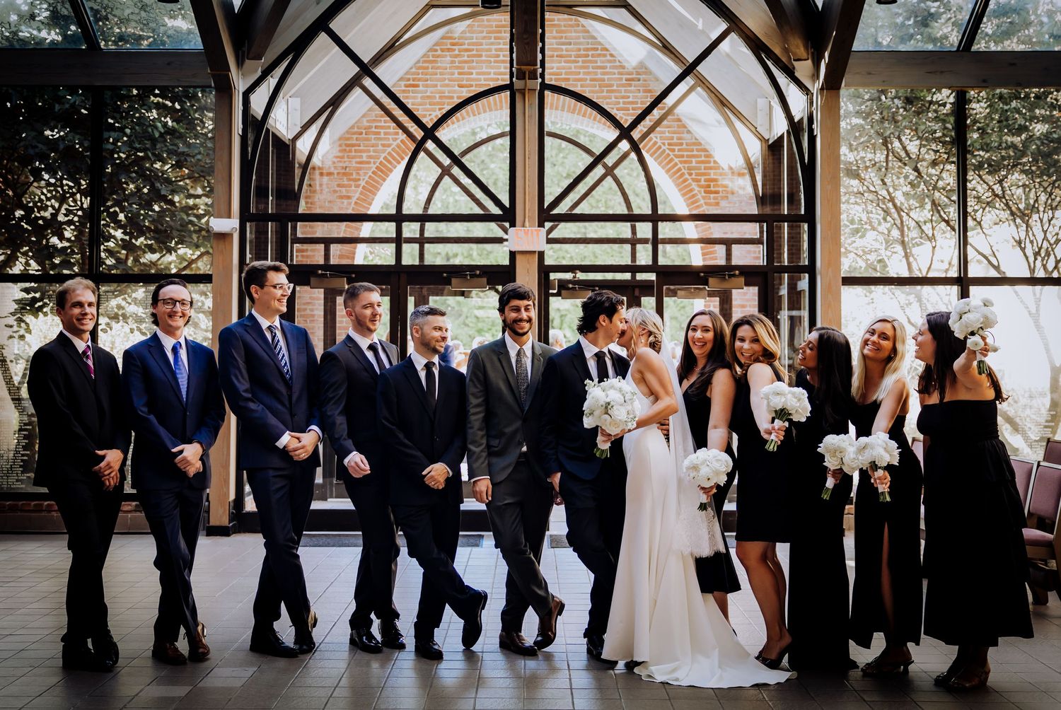 Wedding party poses in formal attire against an architectural backdrop with arched windows.