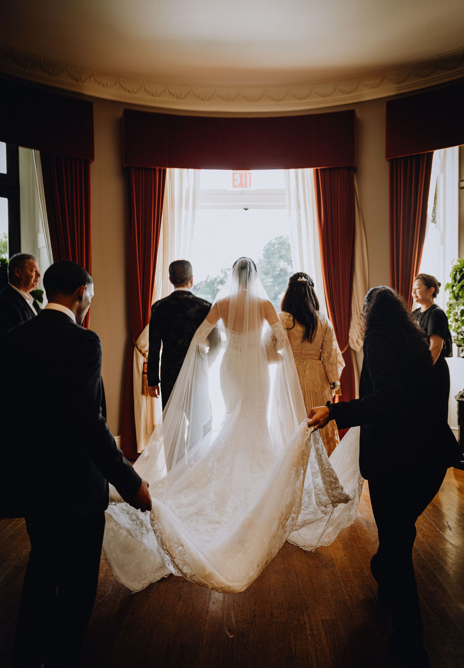 Bride and groom stand before a bright window as attendants arrange the wedding gown train in an elegant venue.