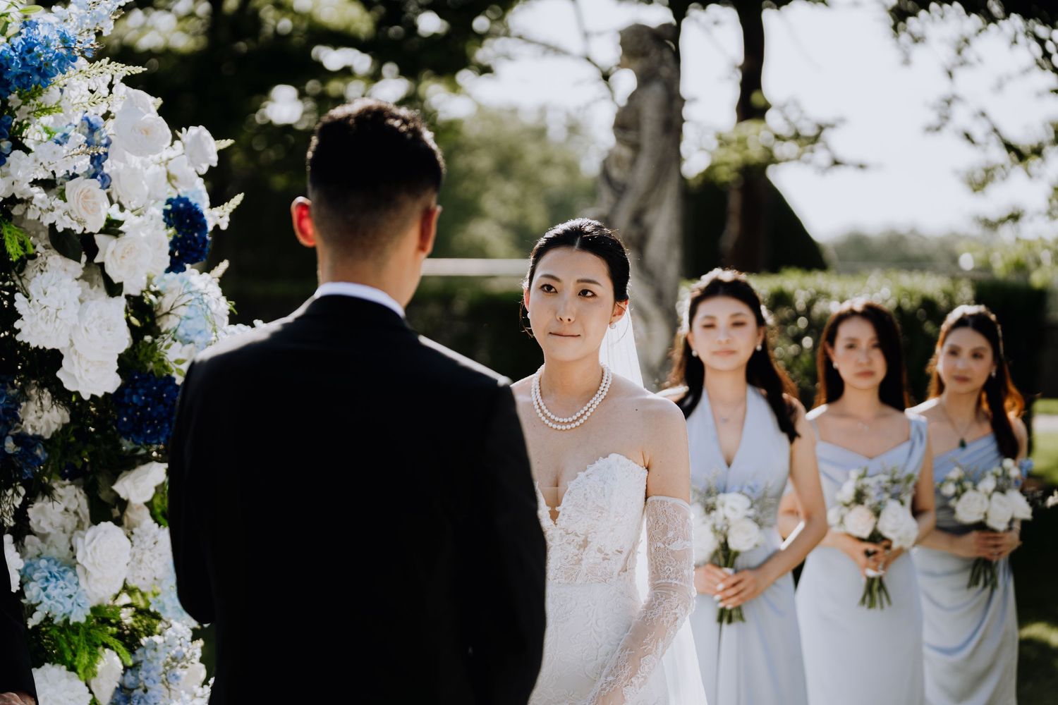 Bride in lace gown with pearl necklace stands at outdoor wedding altar with bridesmaids in blue gowns holding bouquets.