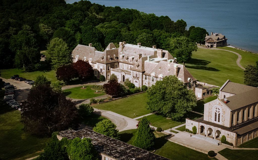 Aerial view of a grand historic stone mansion estate surrounded by lush green trees near a waterfront.