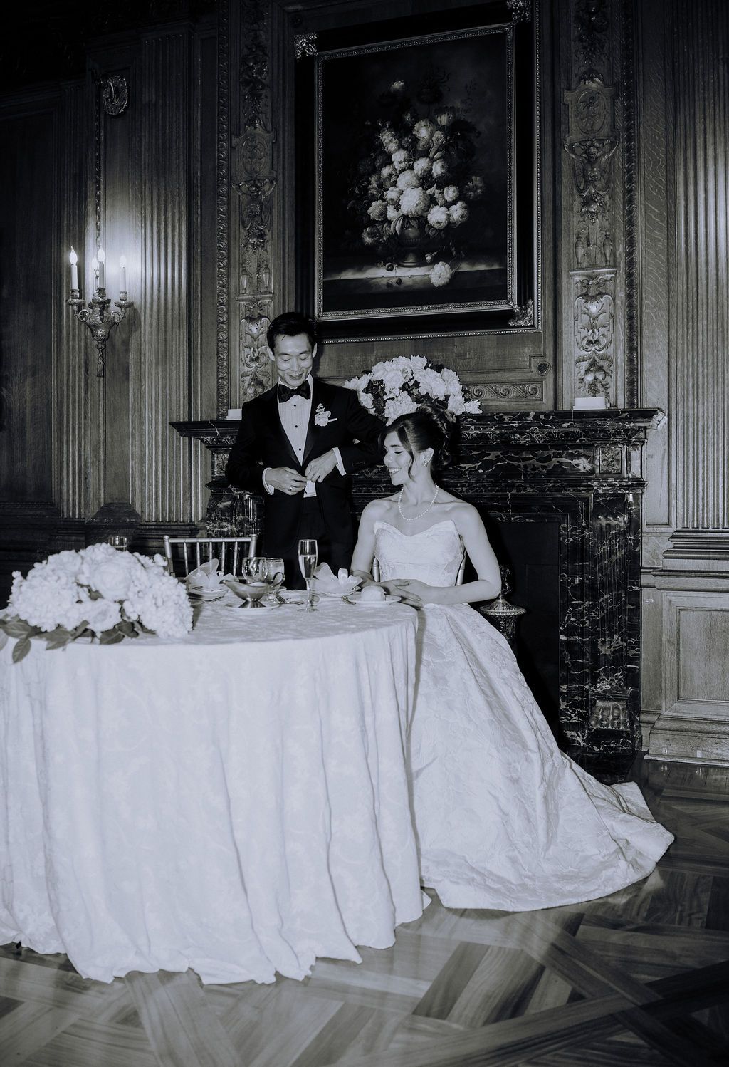 Black and white vintage wedding photo of bride in gown and groom in tuxedo at elegant reception table by fireplace.