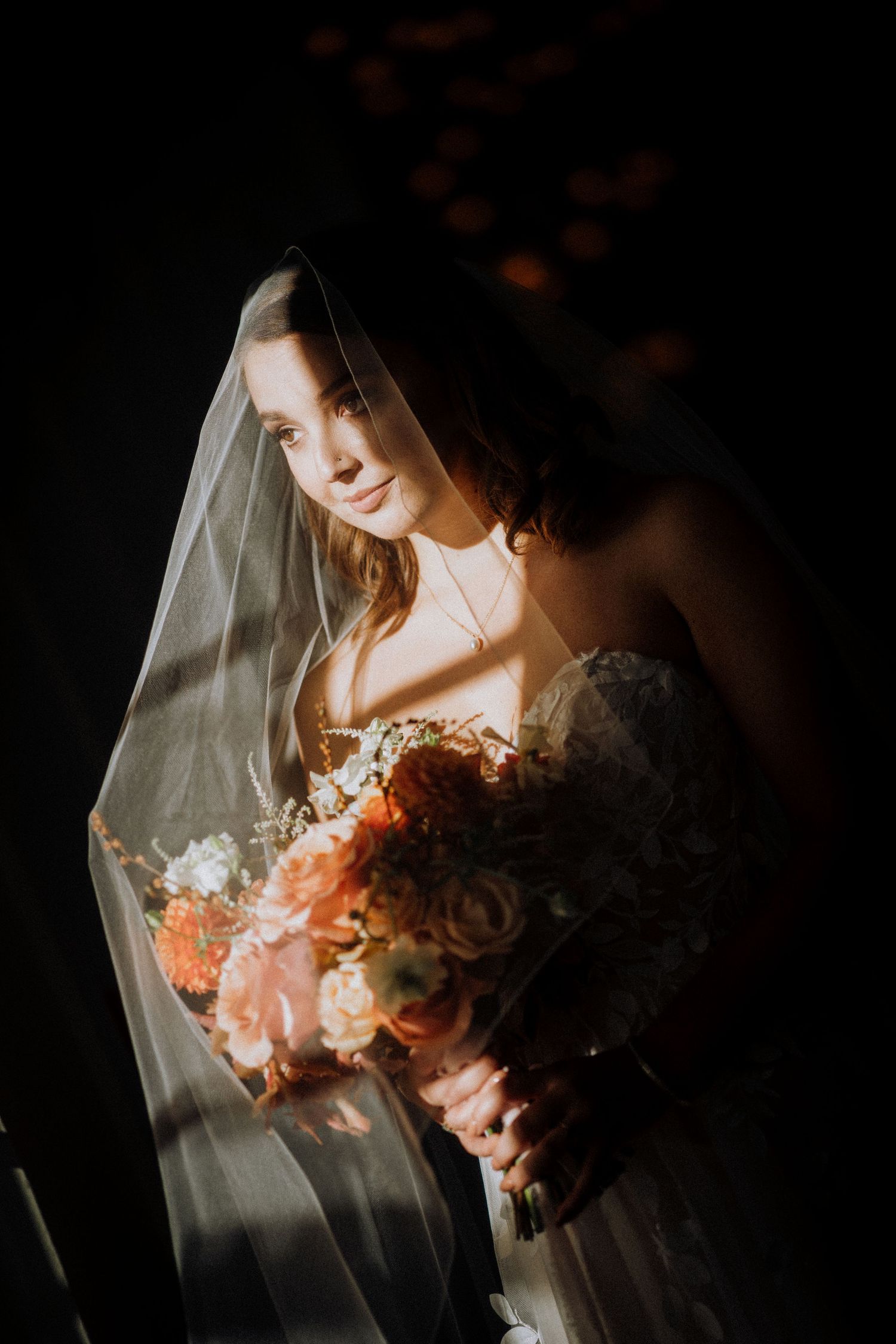 Moody bridal portrait with peach and white bouquet under dramatic lighting and flowing veil.